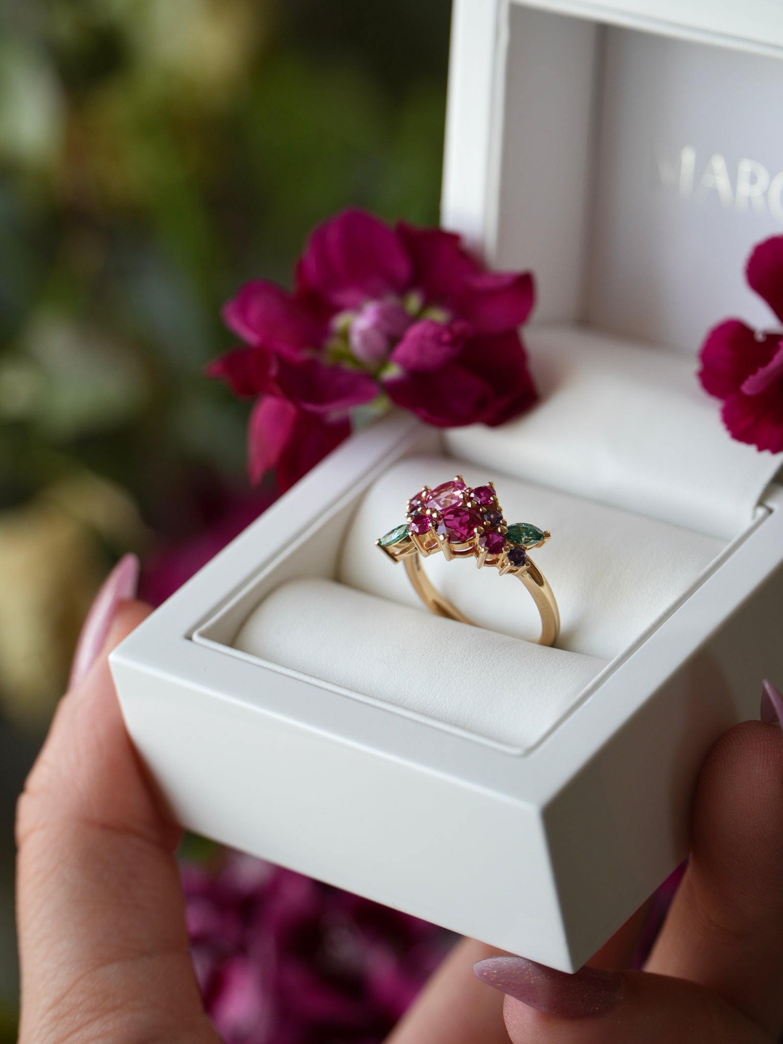 Gold ring with gemstones in a white box held by a hand, with pink flowers in the background