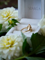 Gold ring in a white box with 'MARGA' branding, surrounded by white flowers.