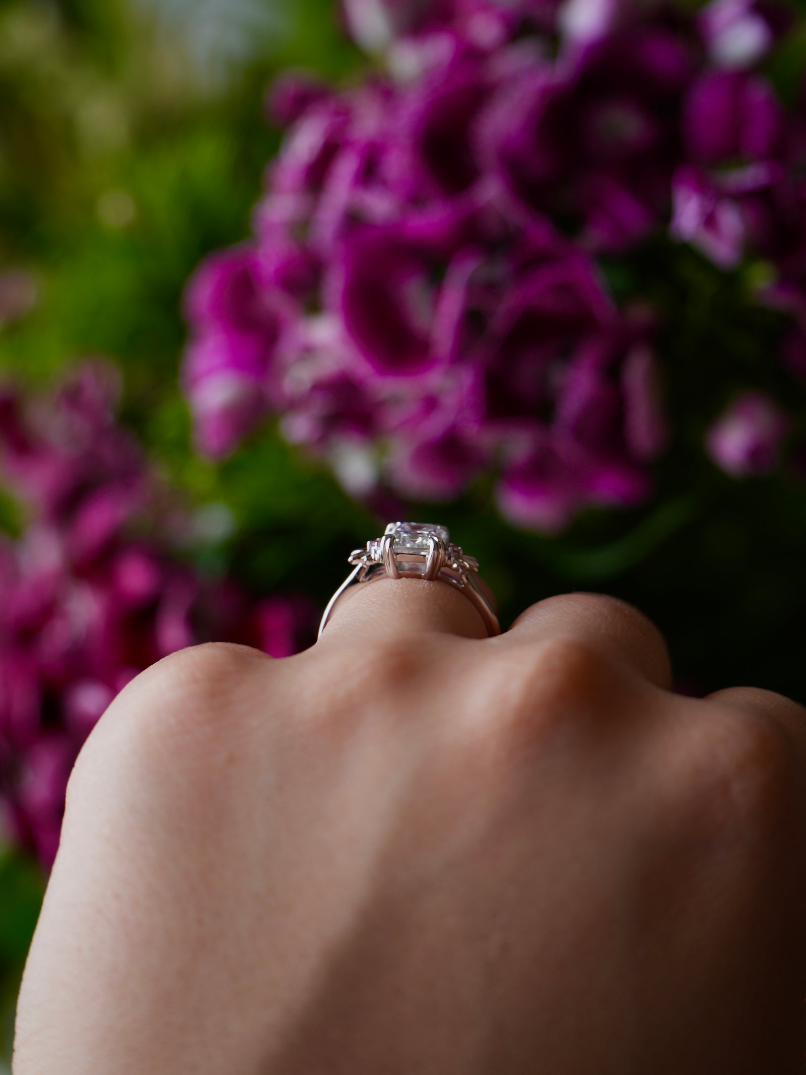 Hand wearing a diamond ring with purple flowers in the background