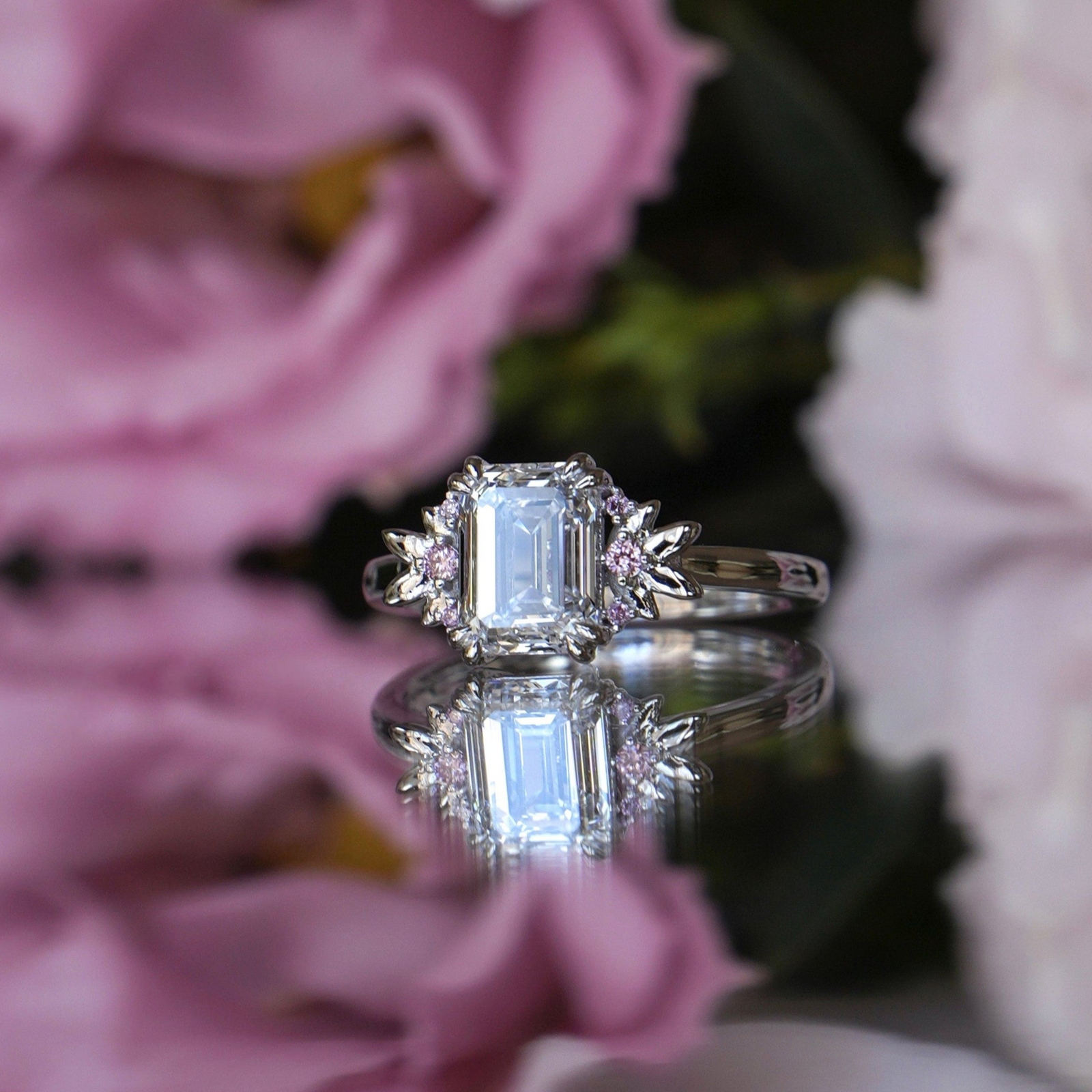 An emerald cut Lab diamond ring with pink round Lab diamonds set in a white gold band, surrounded by white gold leaves, displayed against a floral background.