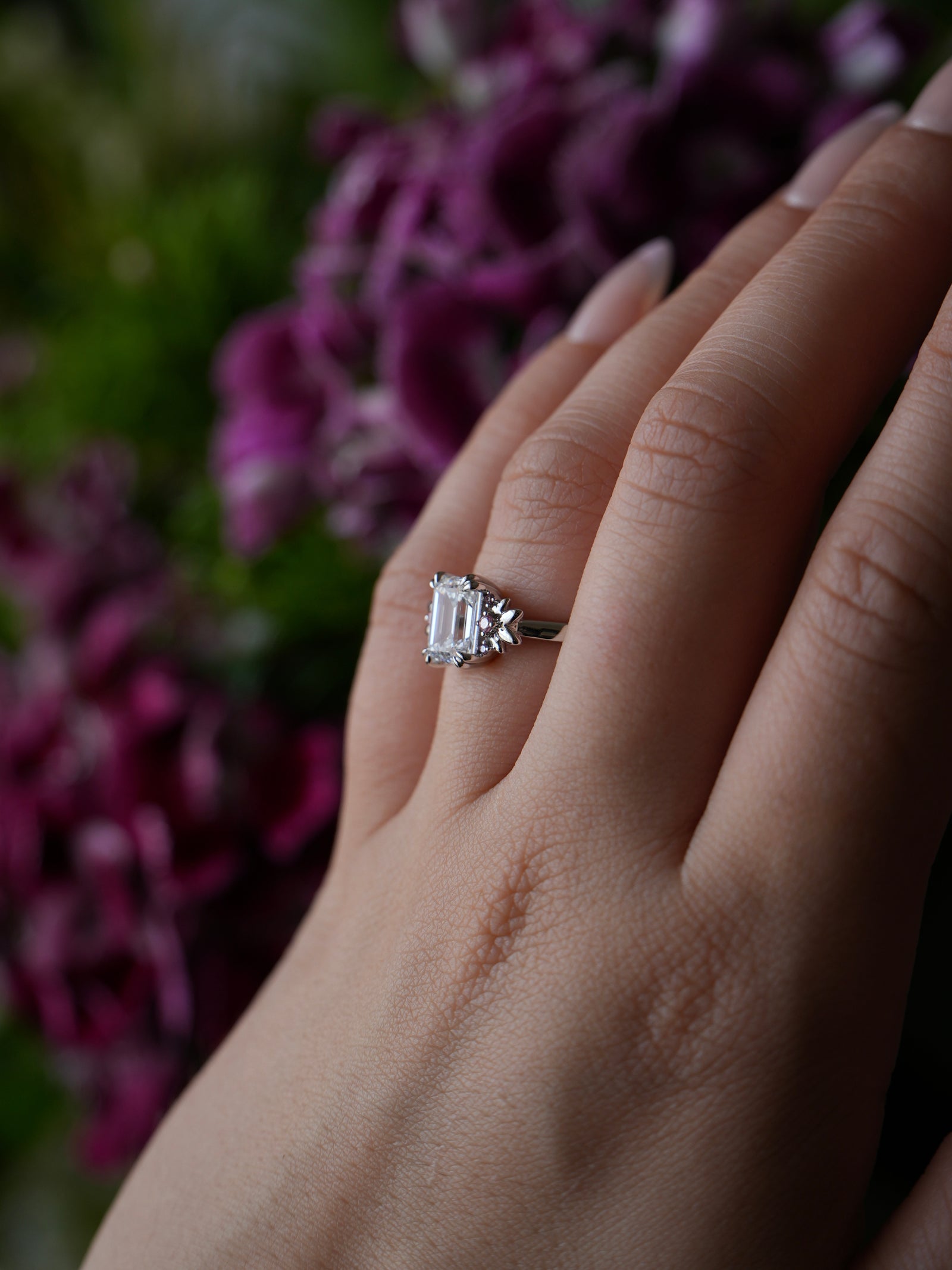 Hand wearing a diamond ring with purple flowers in the background