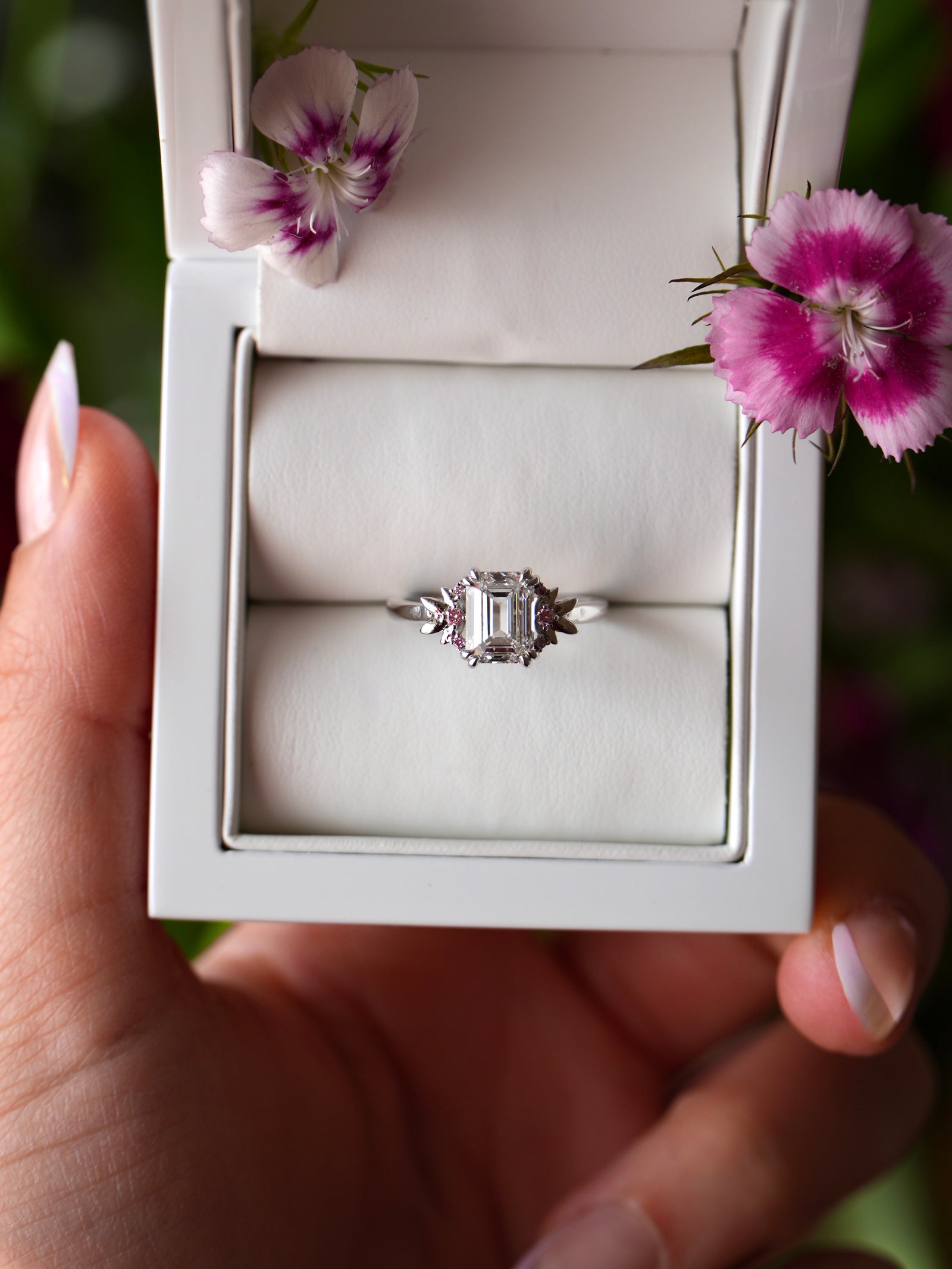 Silver ring with a diamond in a white box held by a hand with pink nail polish, surrounded by pink flowers.