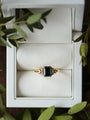 Gold ring with a green gemstone in a white jewelry box surrounded by green leaves.