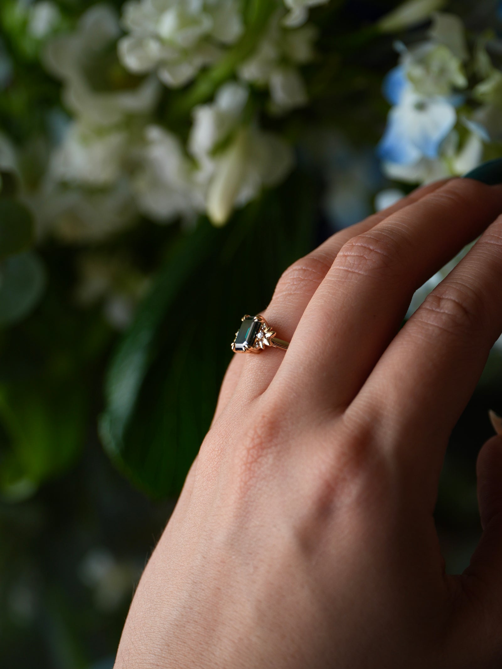 Hand wearing a ring with a green gemstone against a blurred floral background