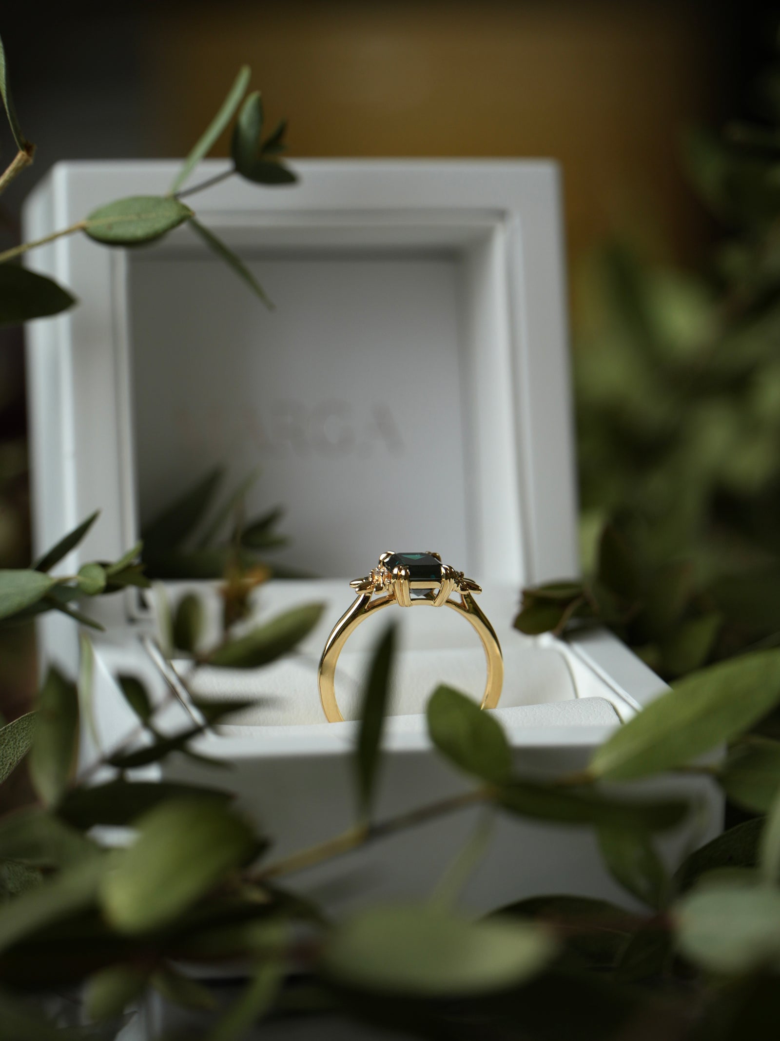 Gold ring with a dark gemstone in a white box surrounded by green leaves
