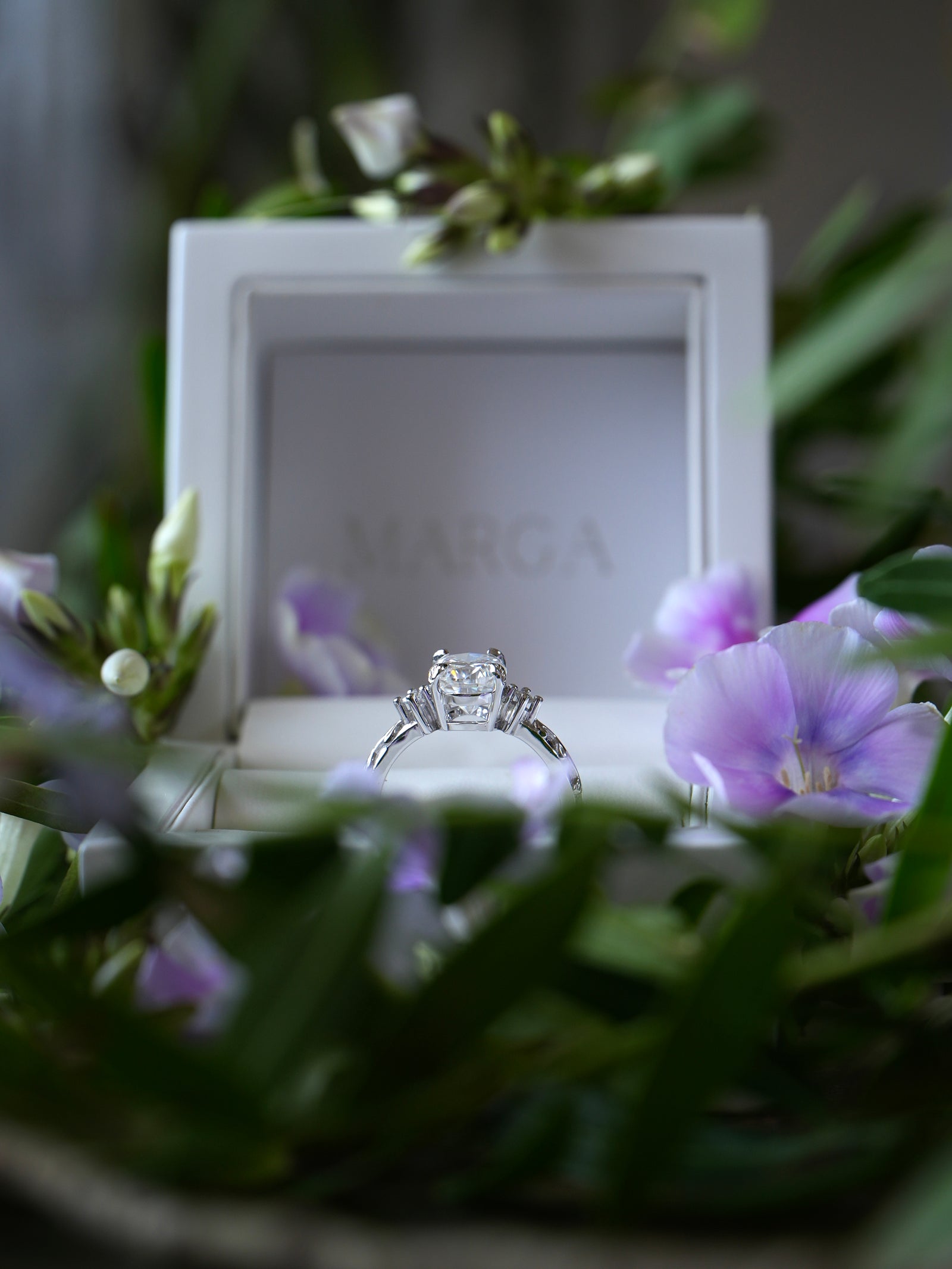 Diamond ring in a white box with 'MARCIA' branding, surrounded by flowers.