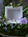 Diamond ring in a white box with 'MARCIA' branding, surrounded by flowers.