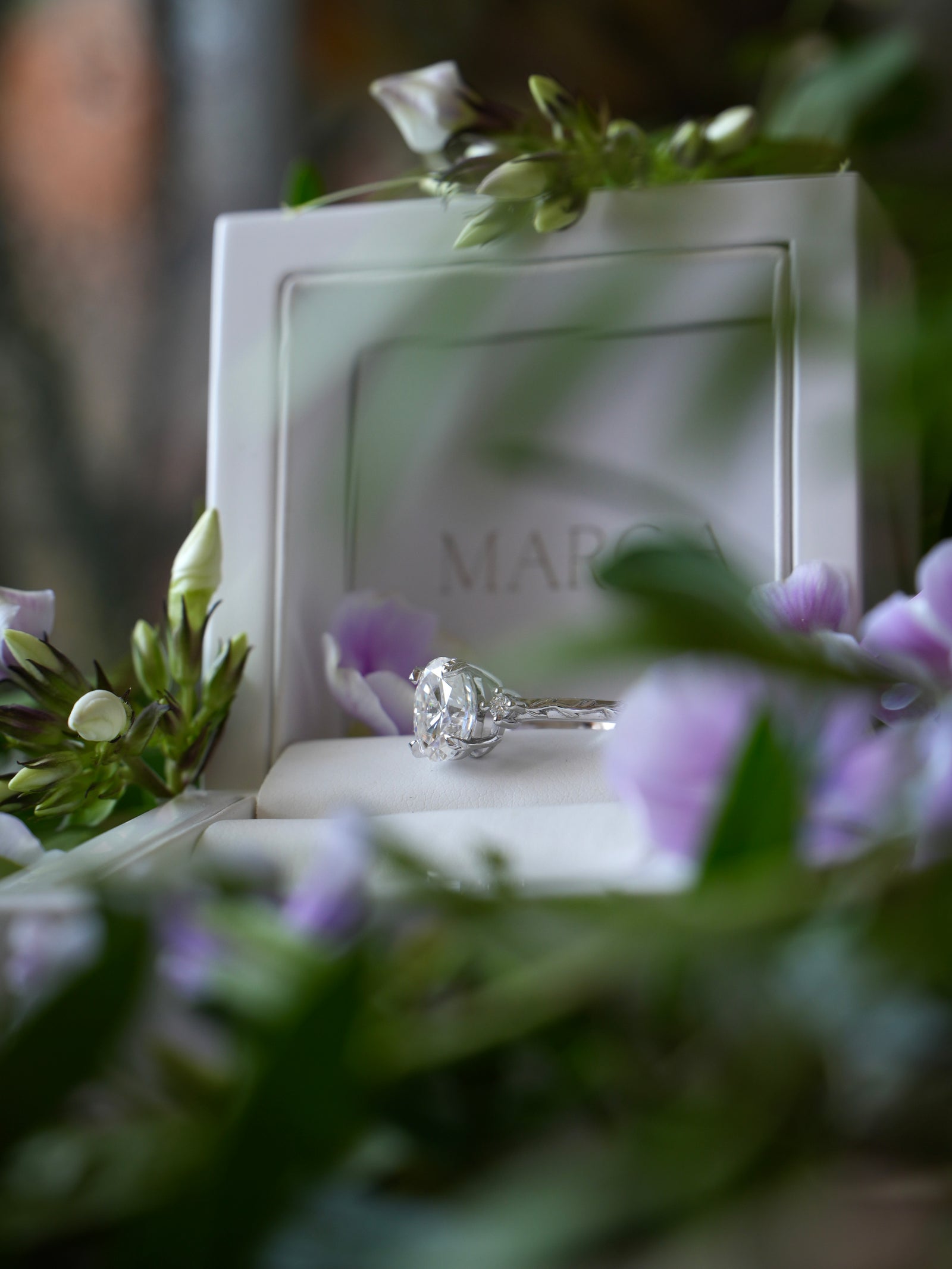Diamond ring in a white box with 'Mare' branding, surrounded by flowers.