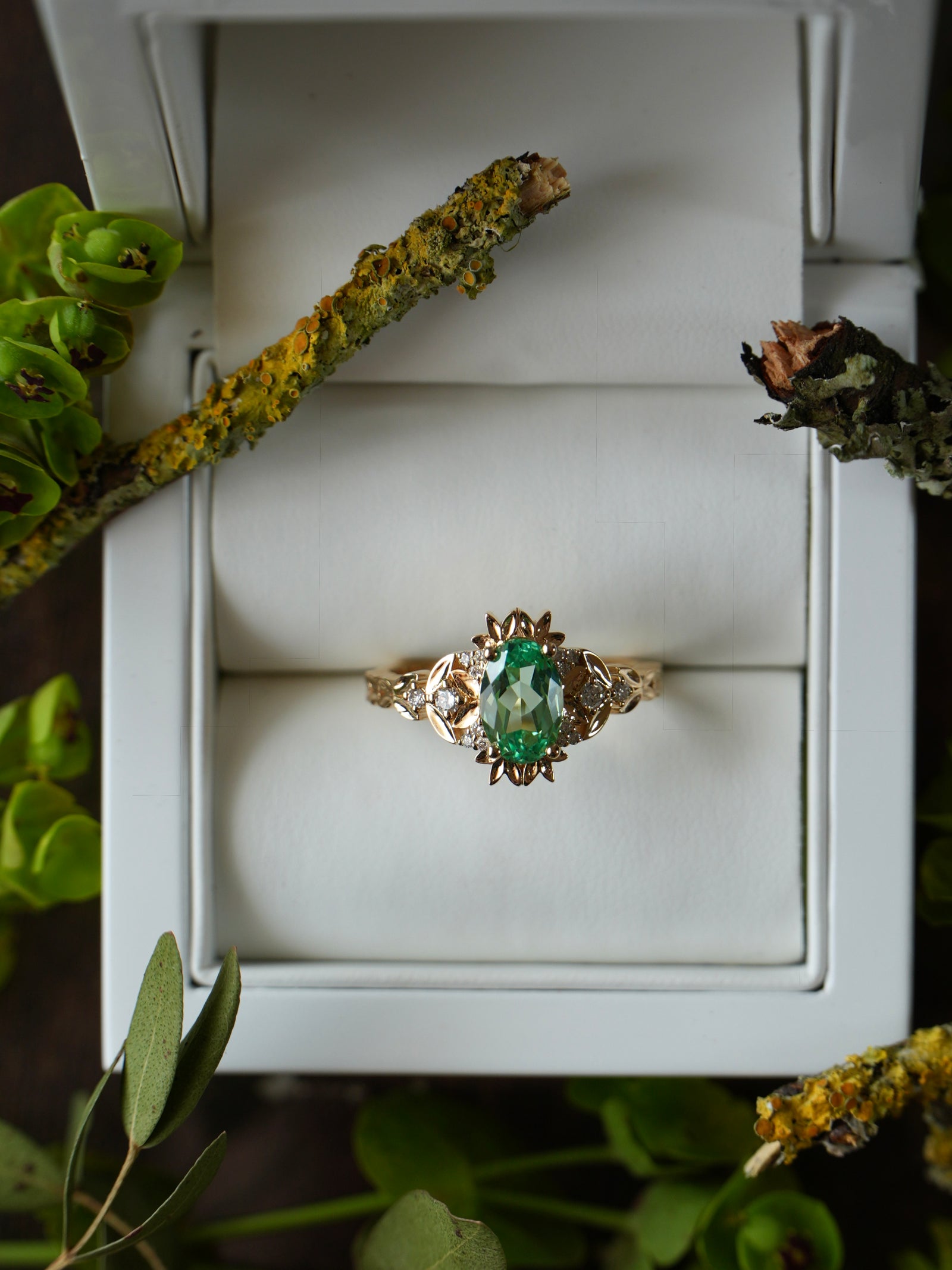 Gold ring with a green gemstone in a white box surrounded by greenery