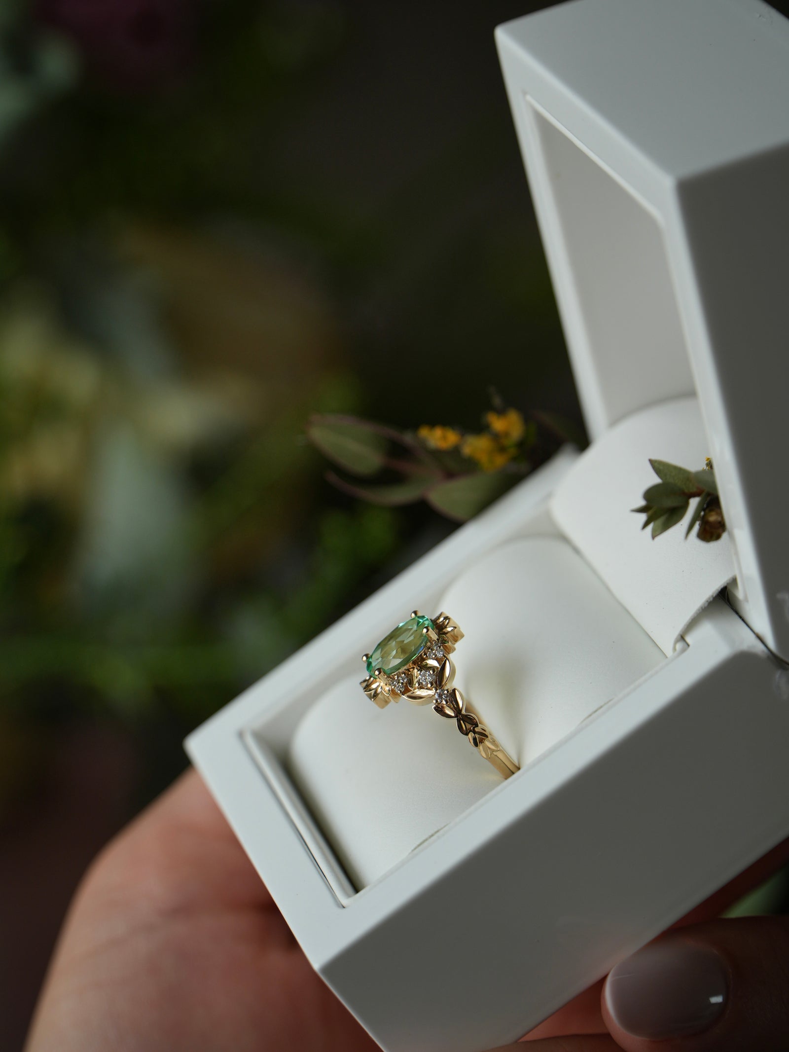Gold ring with a green gemstone in a white jewelry box held by a hand.