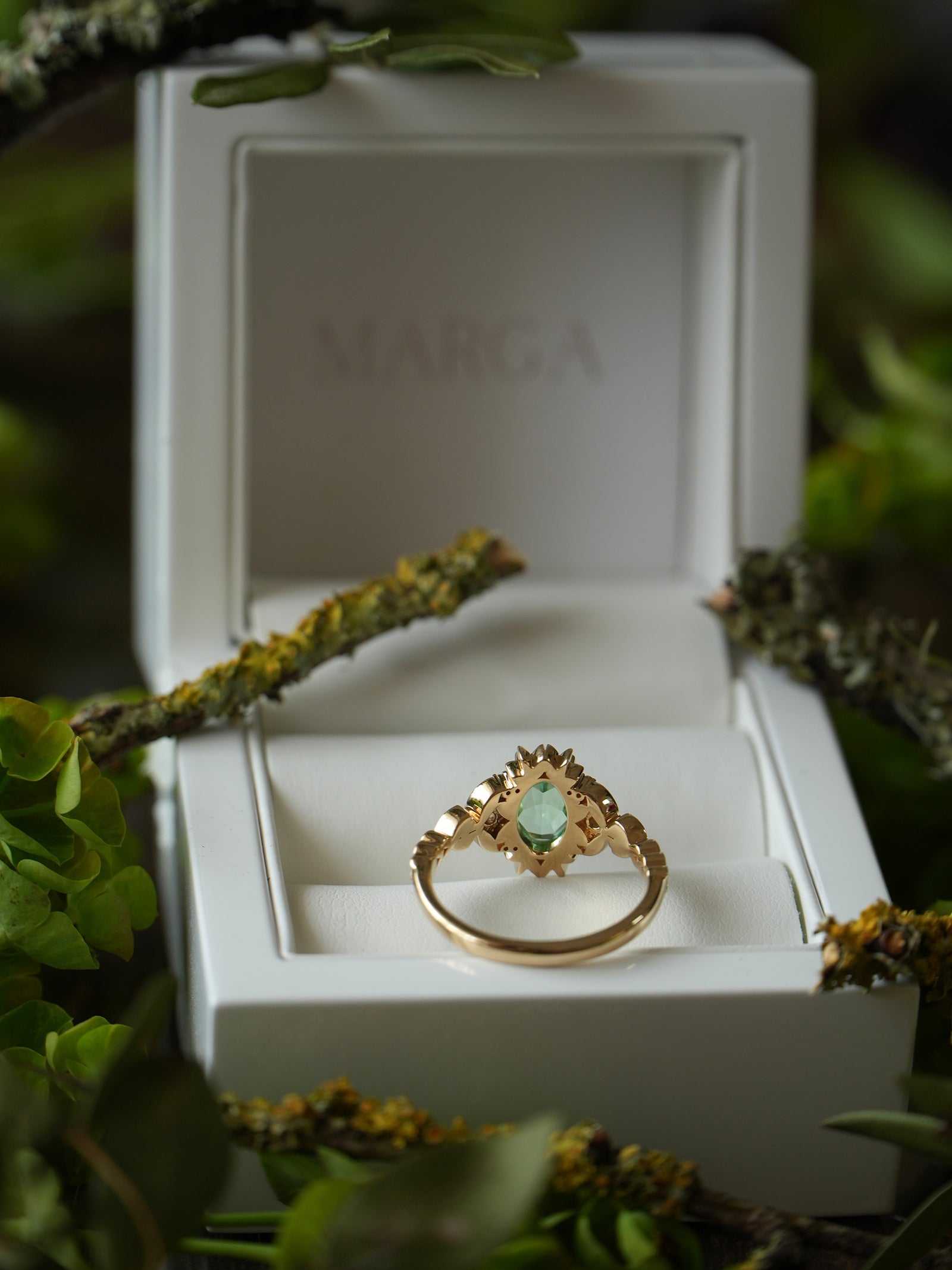 Gold ring with a green gemstone in a white box labeled 'MARGA' surrounded by greenery.