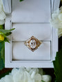 Gold ring with a large diamond in a white box, surrounded by white flowers.