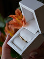 Gold ring in an open white jewelry box held by a hand with a blurred floral background