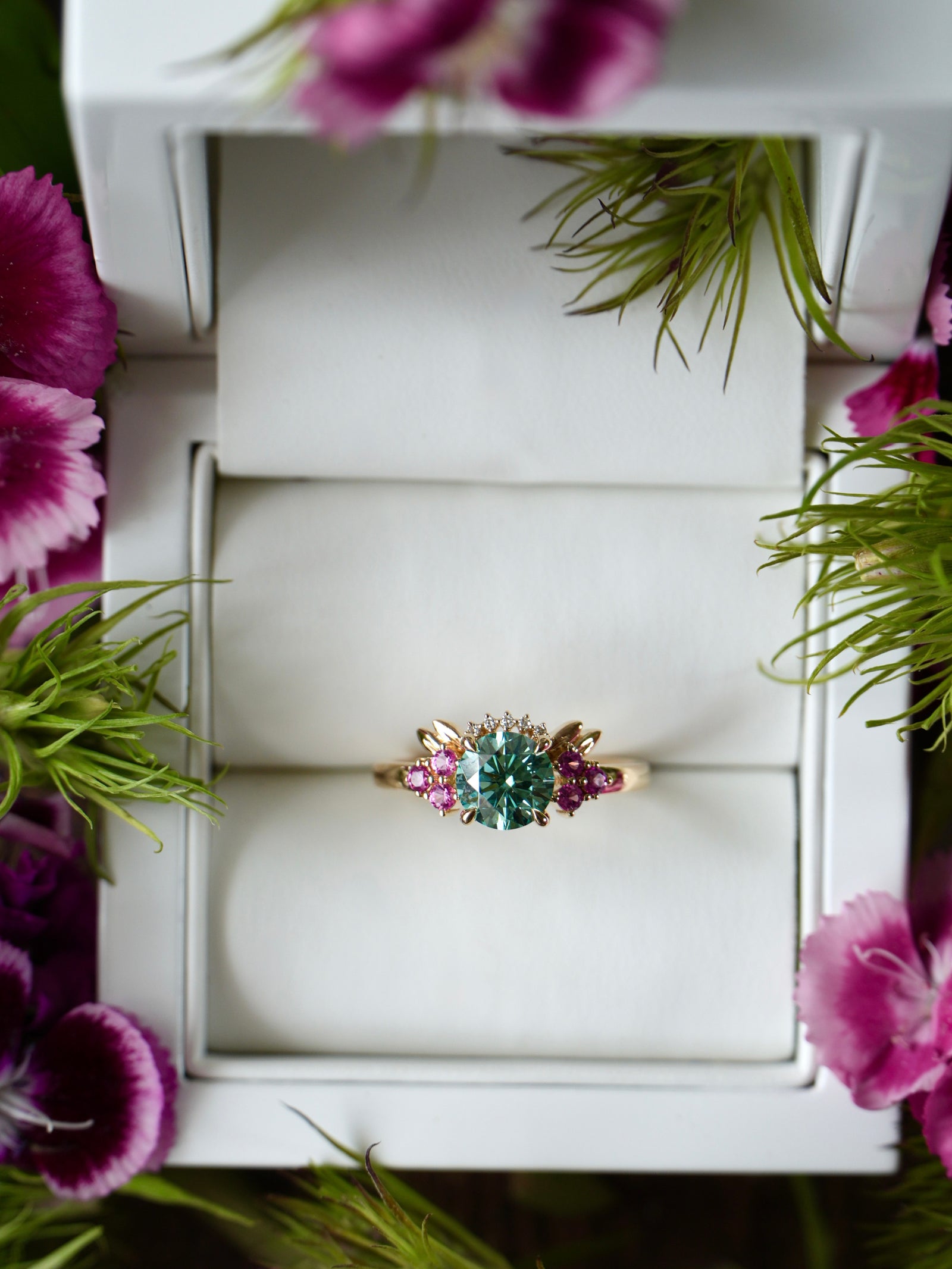 Ring with gemstones in a jewelry box surrounded by flowers and greenery