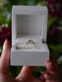 Gold ring with a green gemstone in a white box held by a hand against a blurred natural background