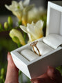 Gold ring with a diamond in a white box held by a hand, with flowers in the background
