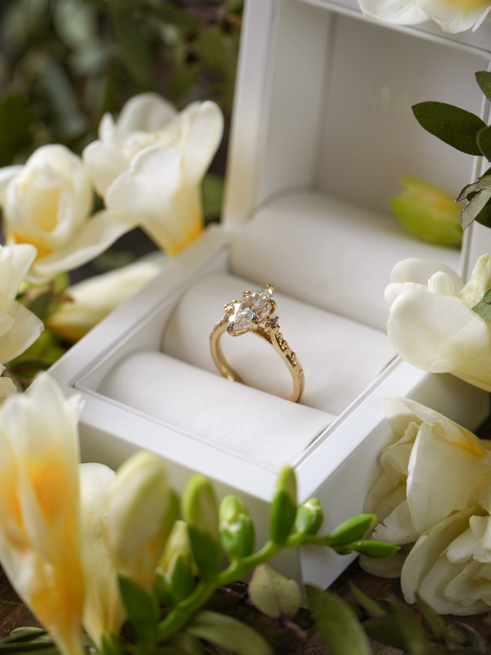 Gold ring with a diamond in a white jewelry box surrounded by white flowers.