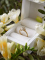 Gold ring with a diamond in a white jewelry box surrounded by white flowers.