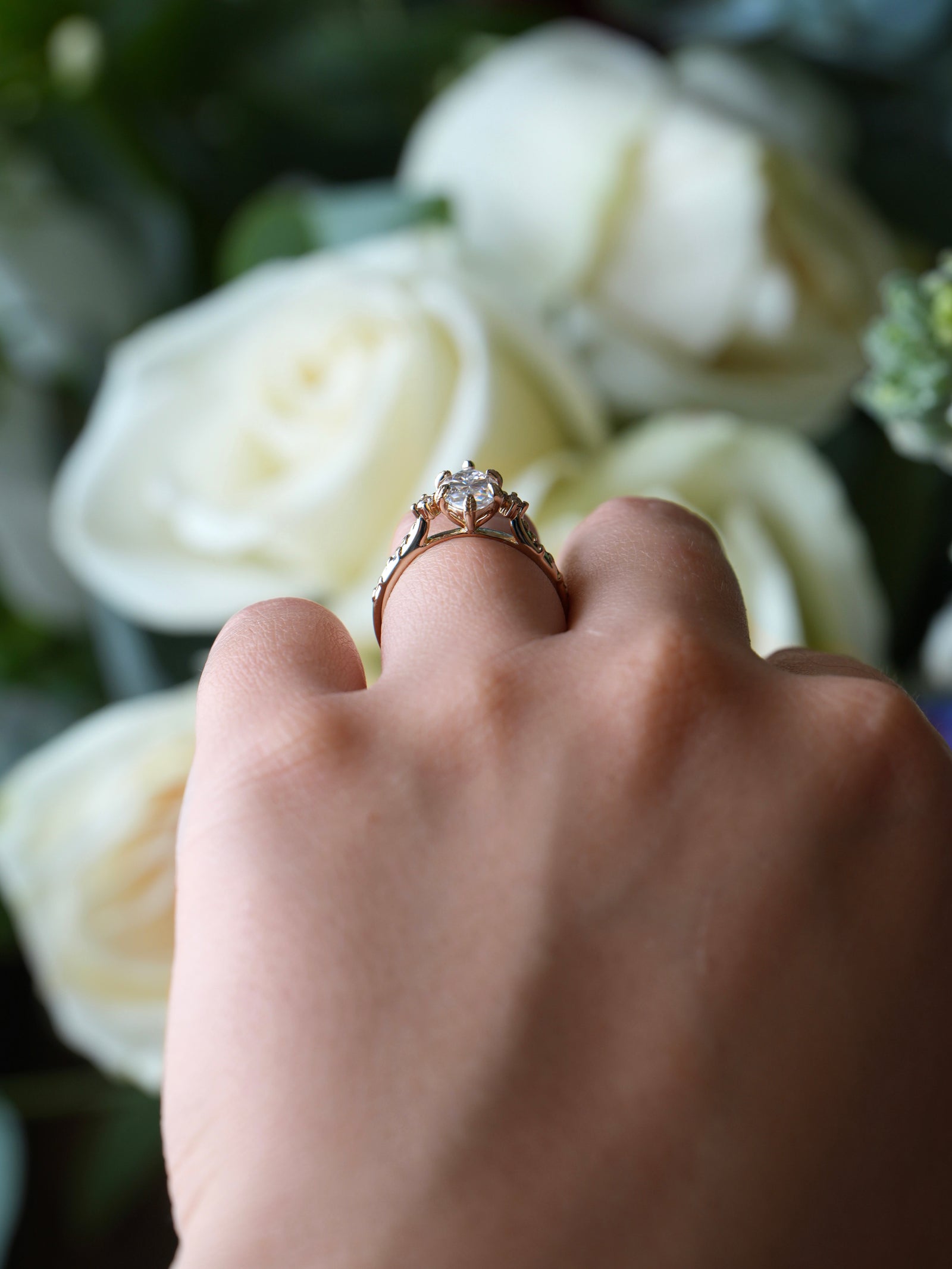 Hand wearing a ring with a diamond, held against a background of white flowers.