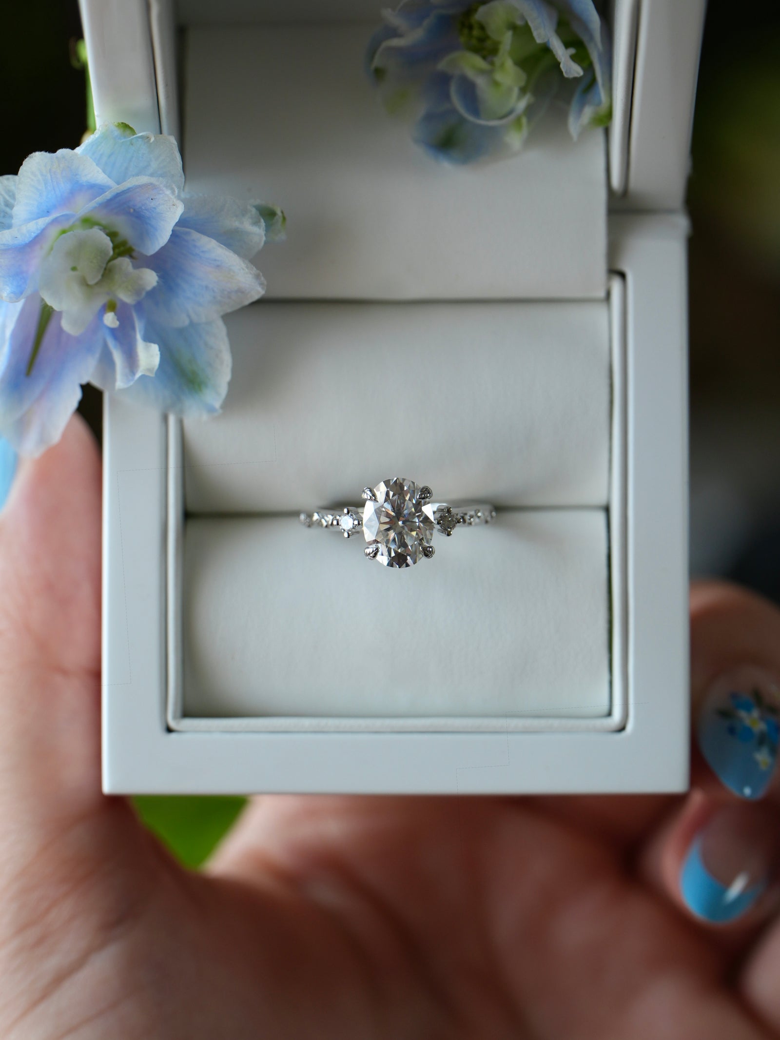 Diamond ring in a white box held by a hand with blue nail polish, surrounded by blue flowers.