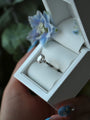 Diamond ring in a white jewelry box held by a hand with floral nail polish.
