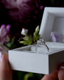 Diamond ring in a white box held by hands with a blurred floral background