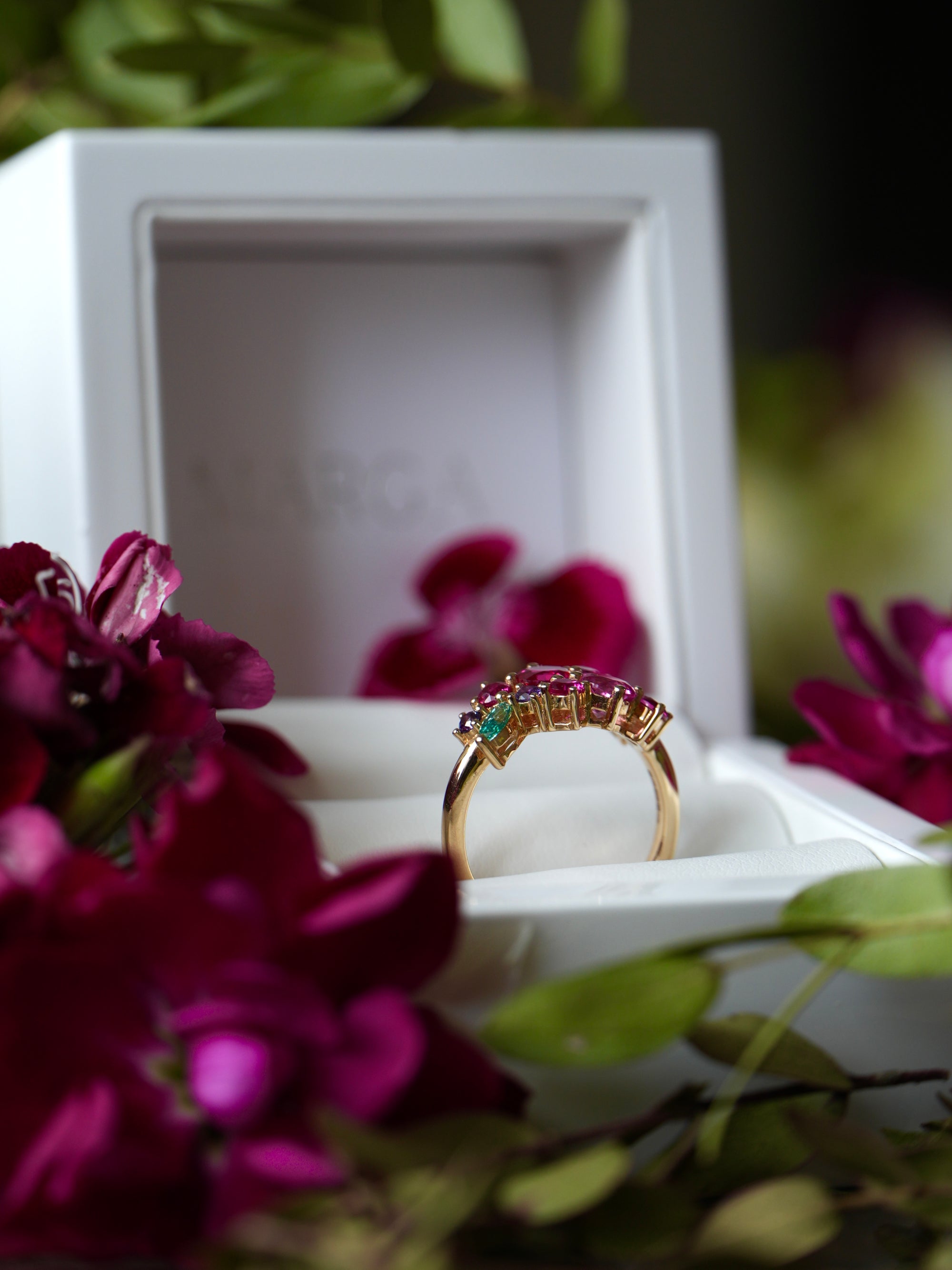 Gold ring with gemstones in a white box surrounded by purple flowers