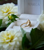 Gold ring in a white box with 'MARGA' branding, surrounded by white flowers.