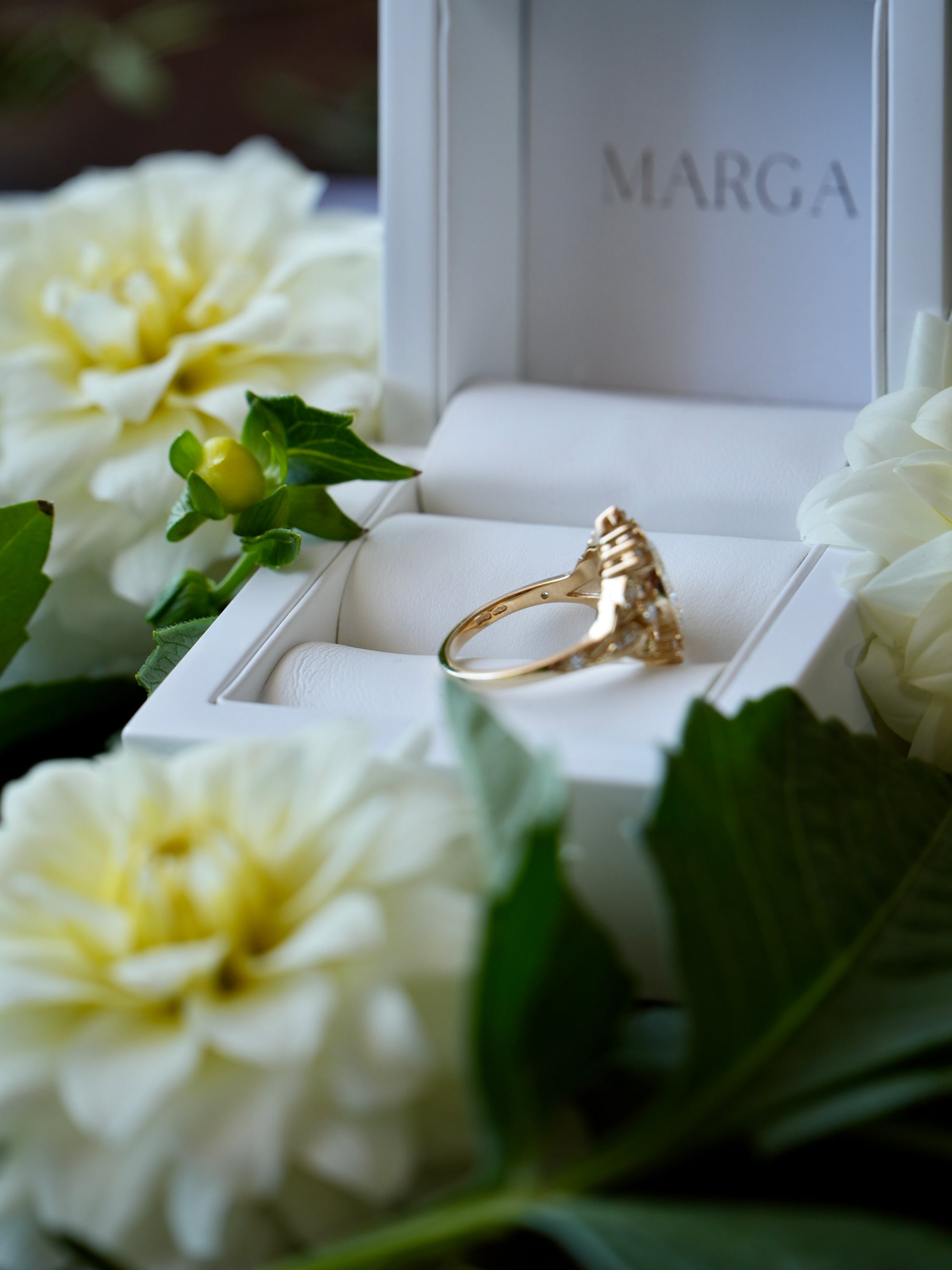 Gold ring in a white box with 'MARGA' branding, surrounded by white flowers.