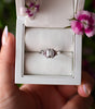 Silver ring with a diamond in a white box held by a hand with pink nail polish, surrounded by pink flowers.