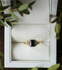 Gold ring with a green gemstone in a white jewelry box surrounded by green leaves.