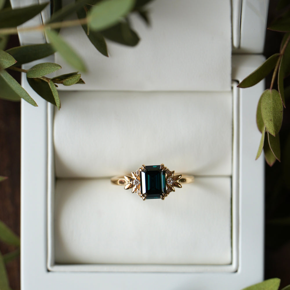 Gold ring with a green gemstone in a white jewelry box surrounded by green leaves.