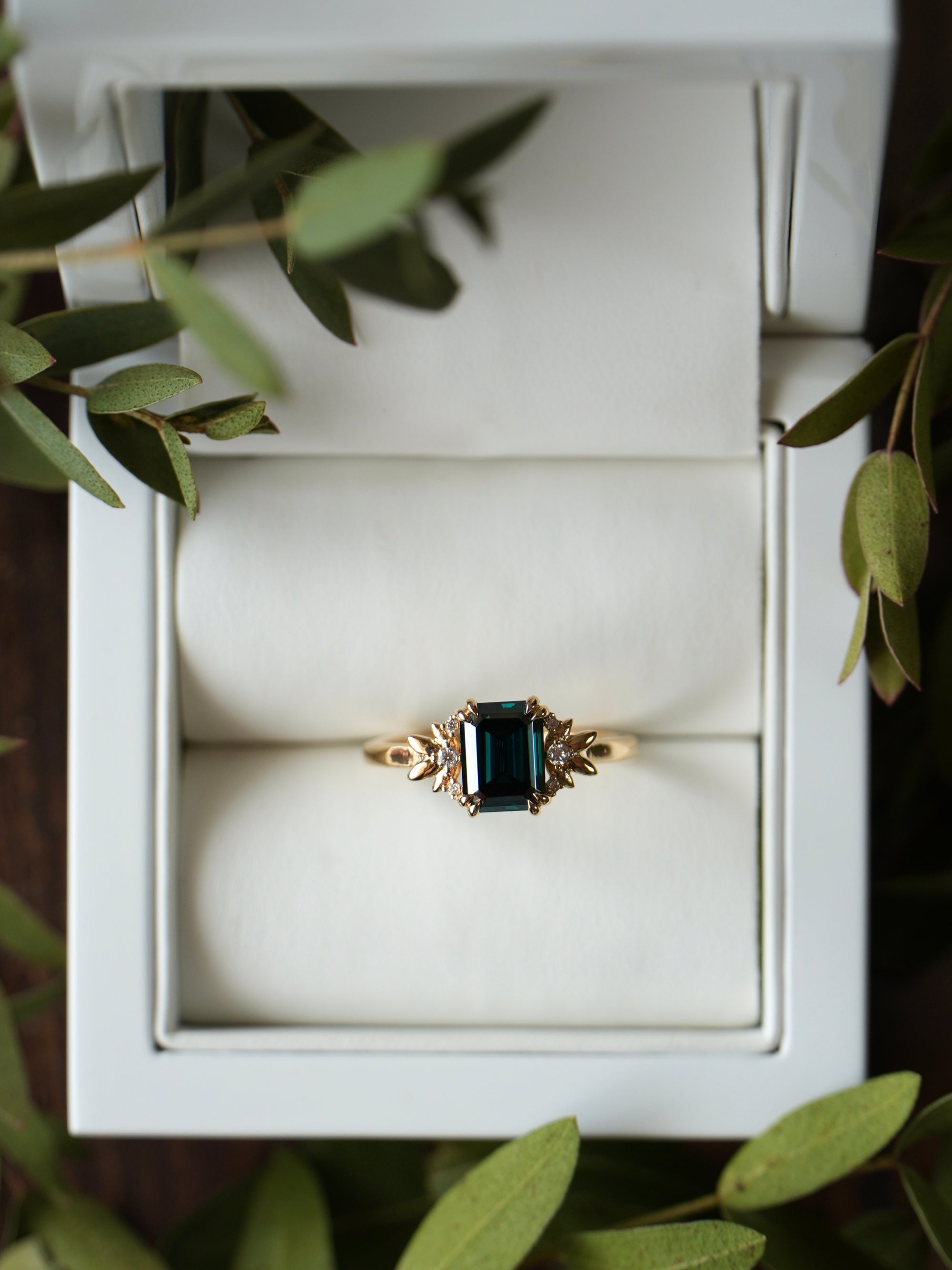 Gold ring with a green gemstone in a white jewelry box surrounded by green leaves.