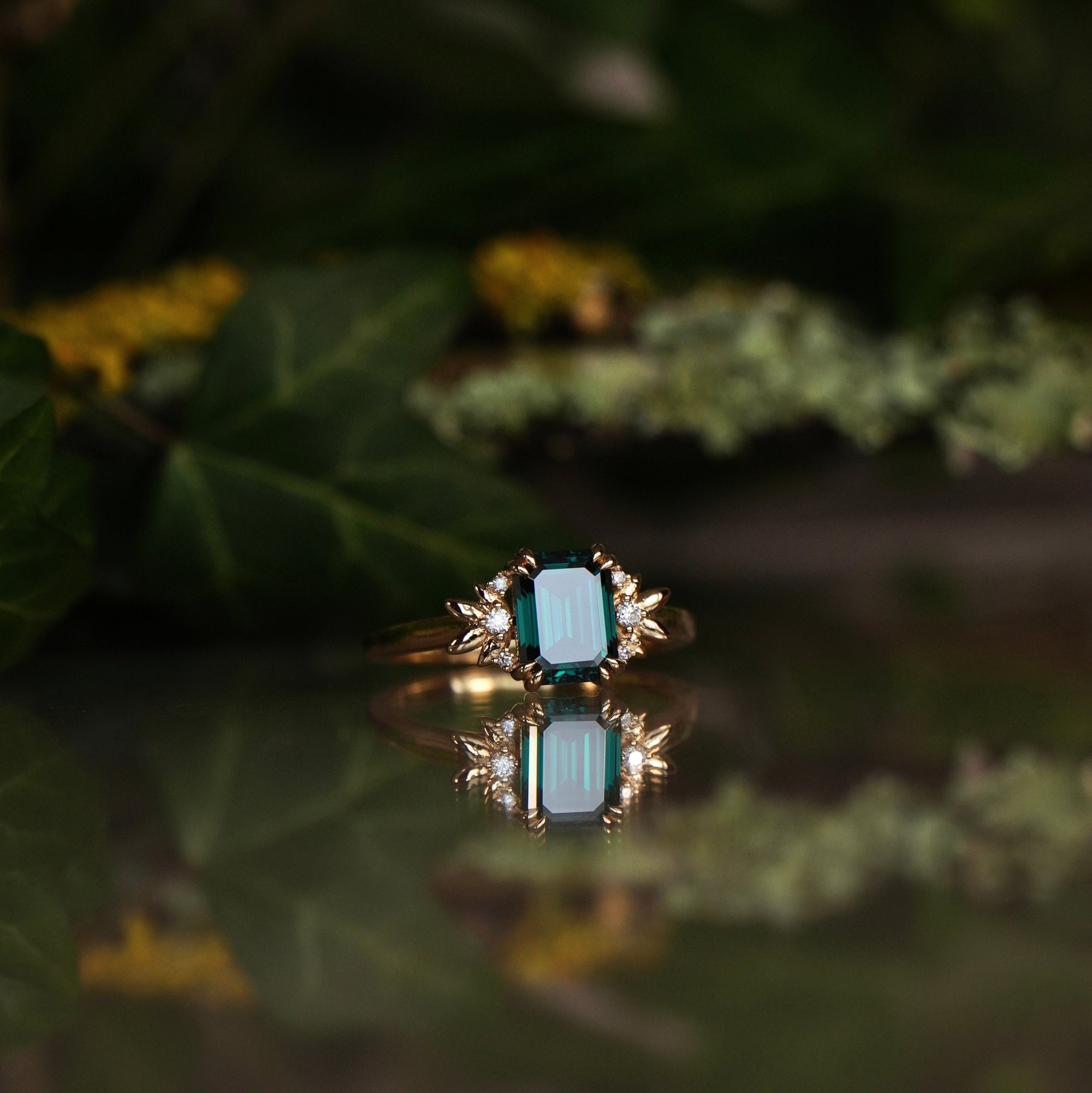 An emerald-cut green moissanite ring with lab diamonds and gold leaves on a reflective surface with a natural background.
