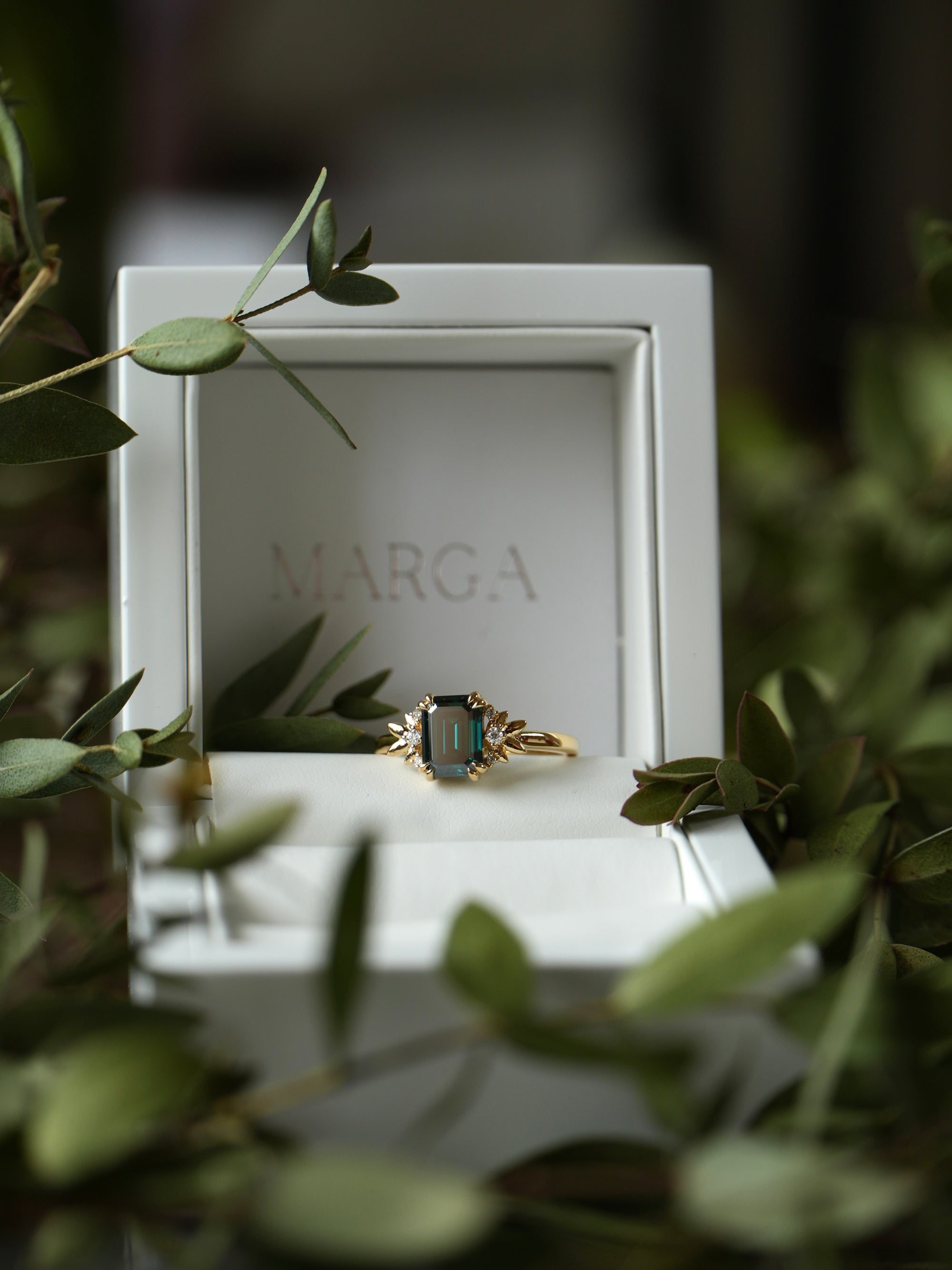 Gold ring with a green gemstone in a MARGA box surrounded by green leaves.