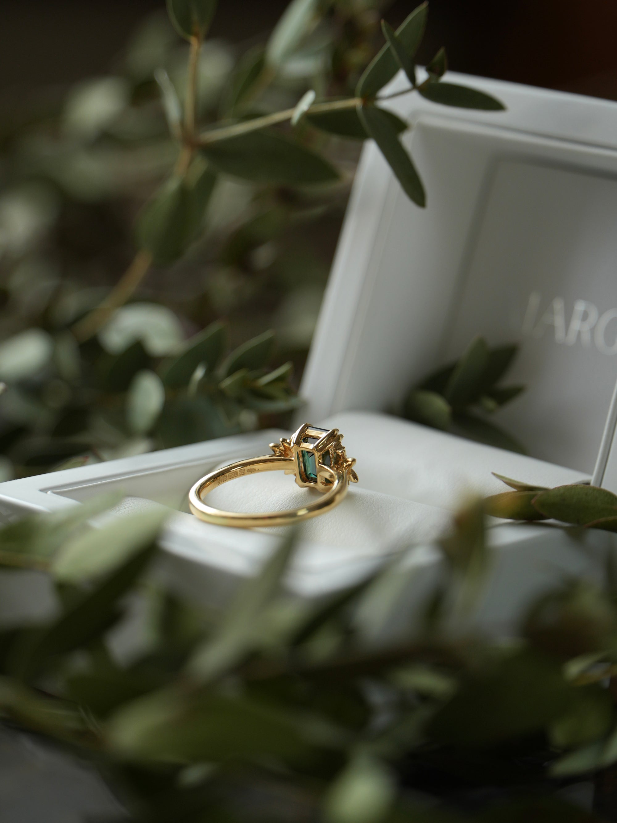 Gold ring with green gemstone on a white surface with blurred green leaves in the foreground