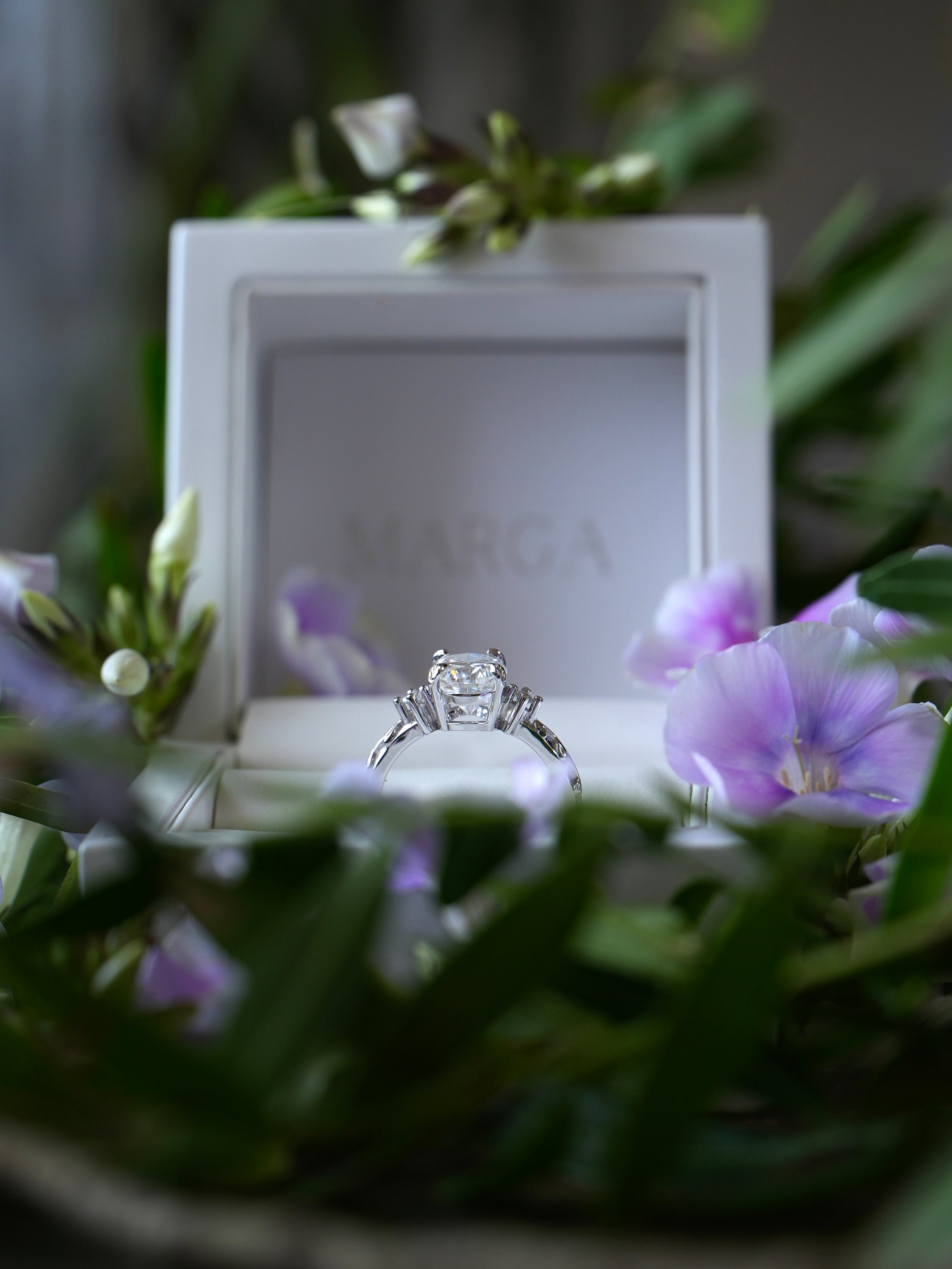 Diamond ring in a white box with 'MARCIA' branding, surrounded by flowers.
