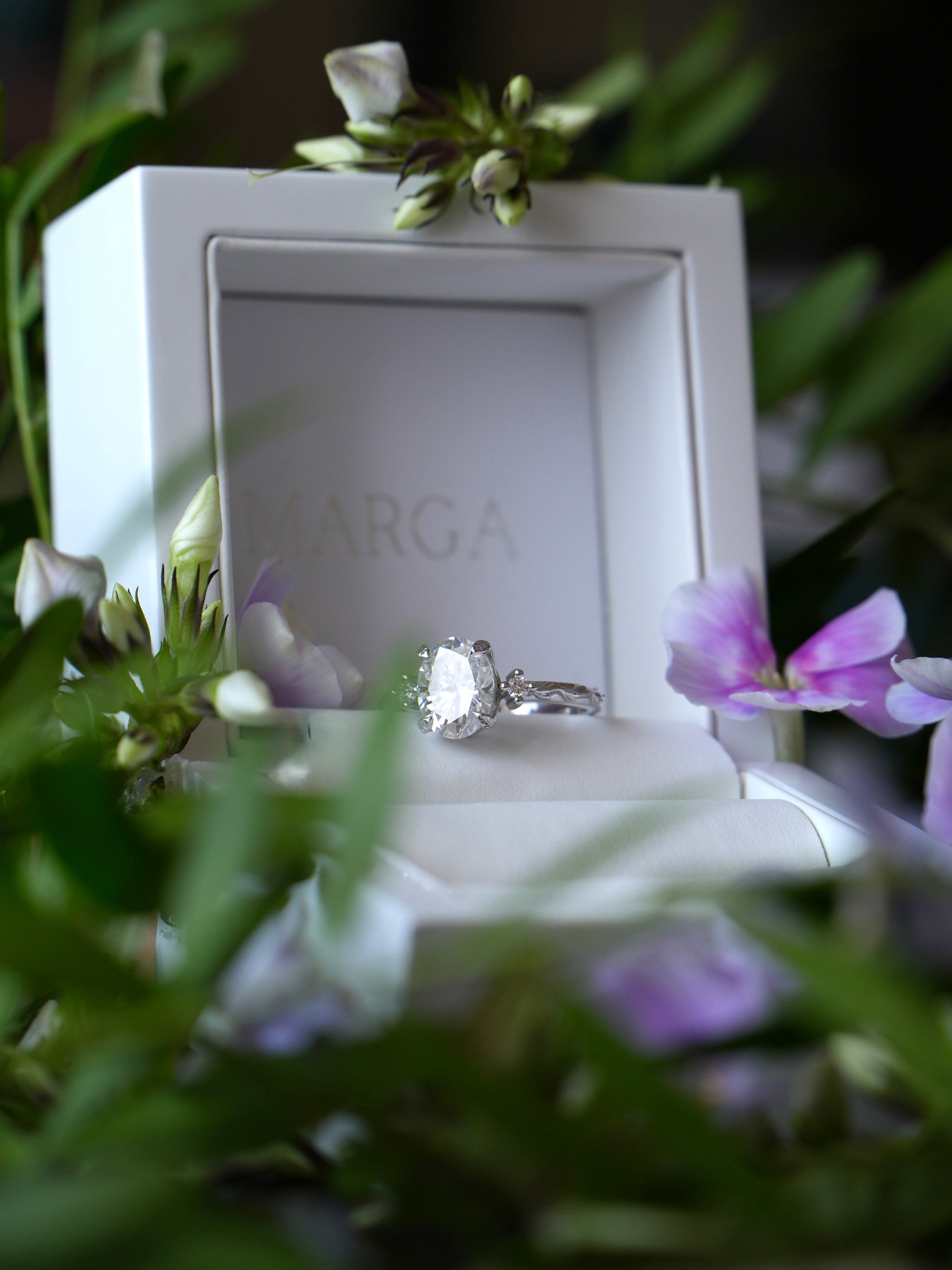 Diamond ring in a white box with 'MARGA' branding, surrounded by flowers.