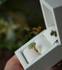 Gold ring with a green gemstone in a white jewelry box held by a hand.
