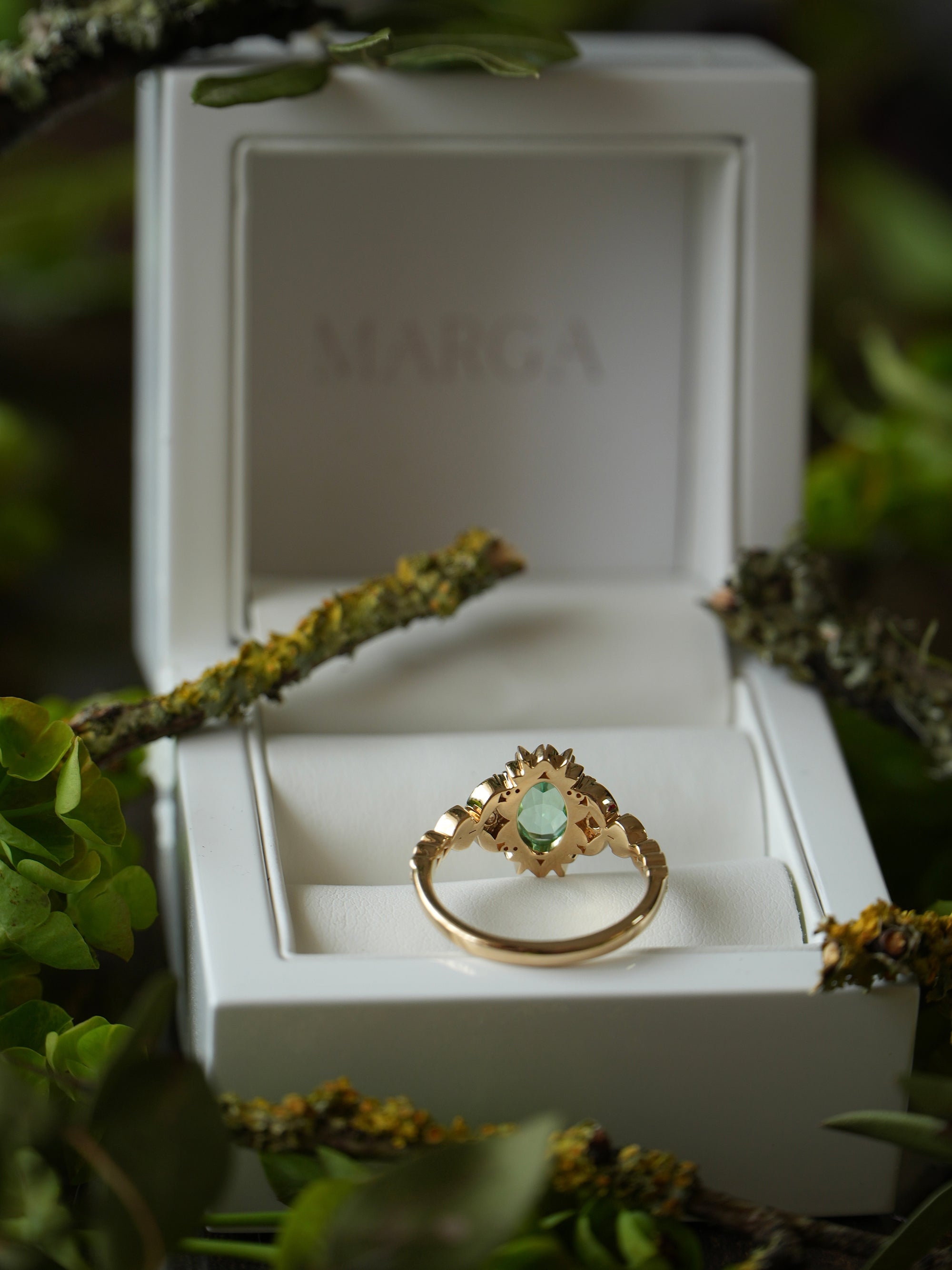Gold ring with a green gemstone in a white box labeled 'MARGA' surrounded by greenery.