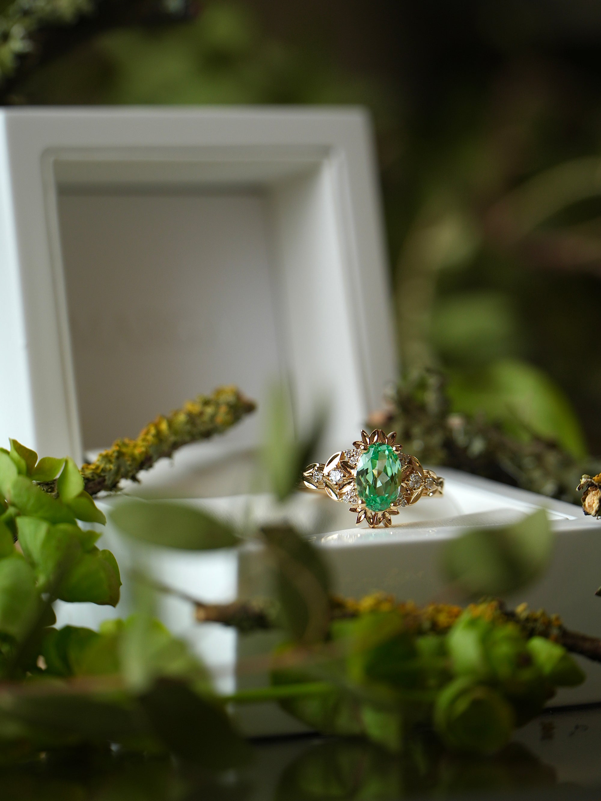 Gold ring with a green gemstone on a white stand surrounded by greenery