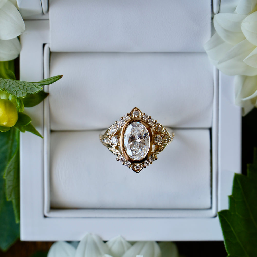Gold ring with a large diamond in a white box, surrounded by white flowers.