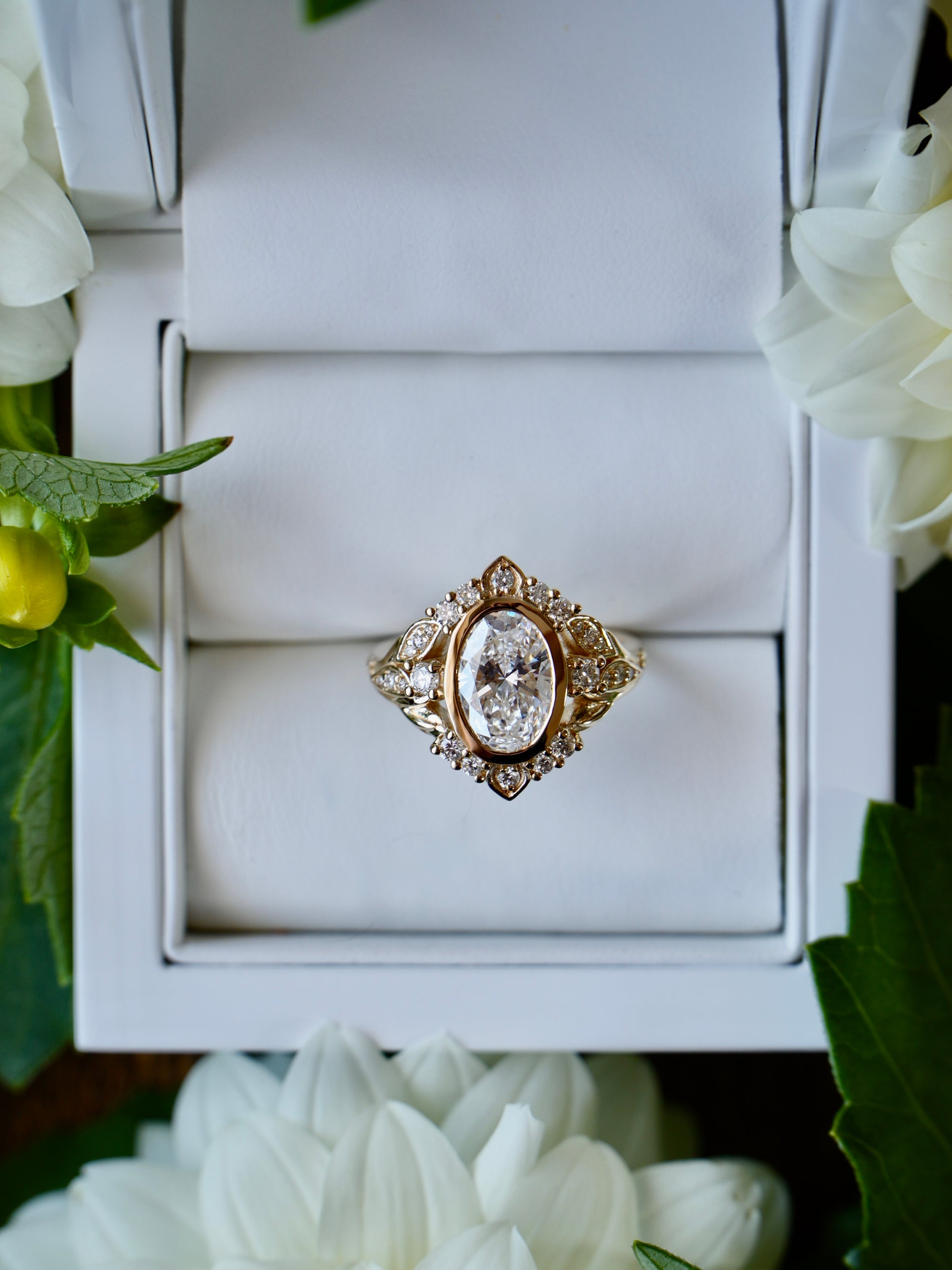 Gold ring with a large diamond in a white box, surrounded by white flowers.