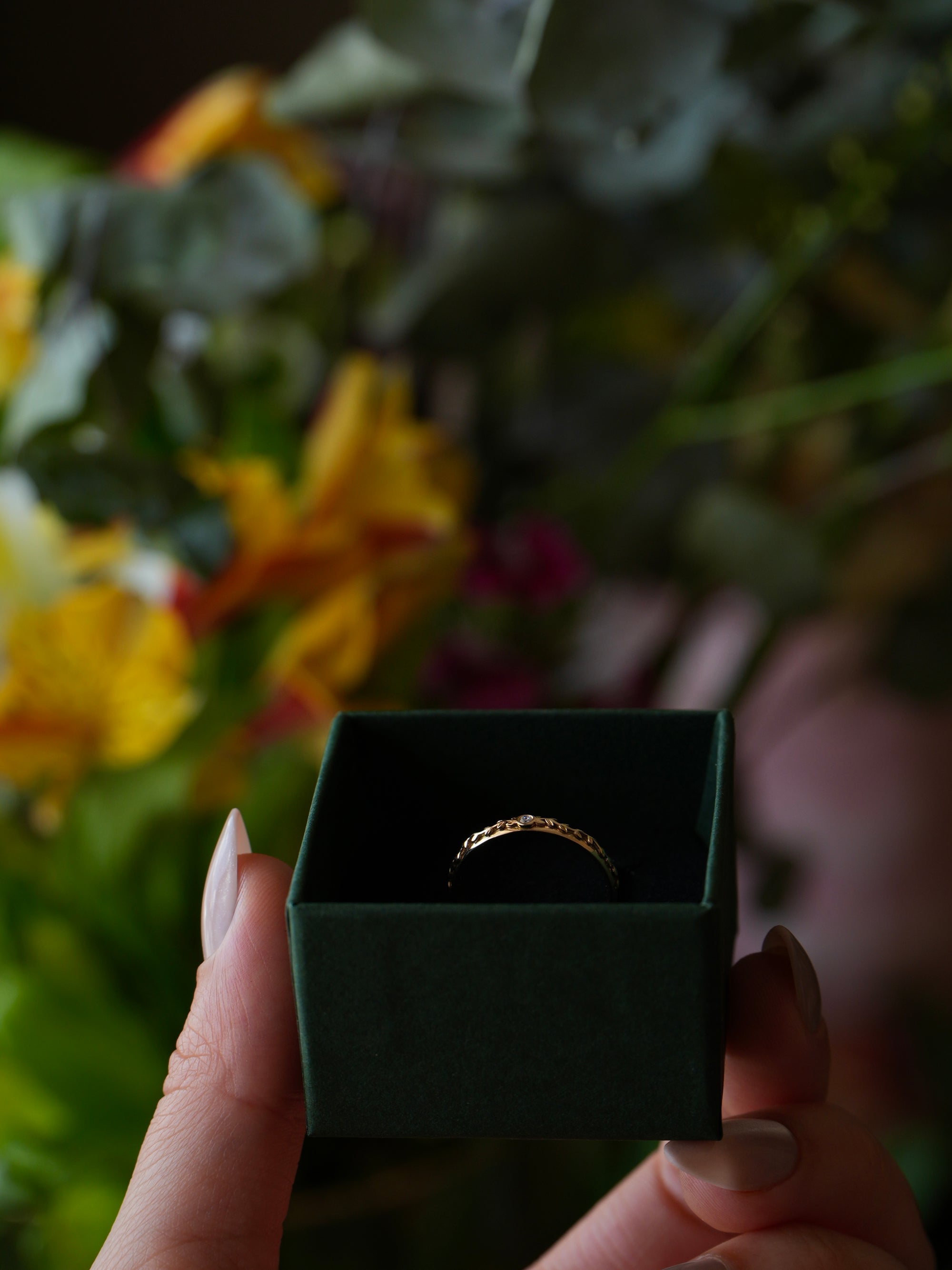 Gold ring in a black box held by a hand with flowers in the background