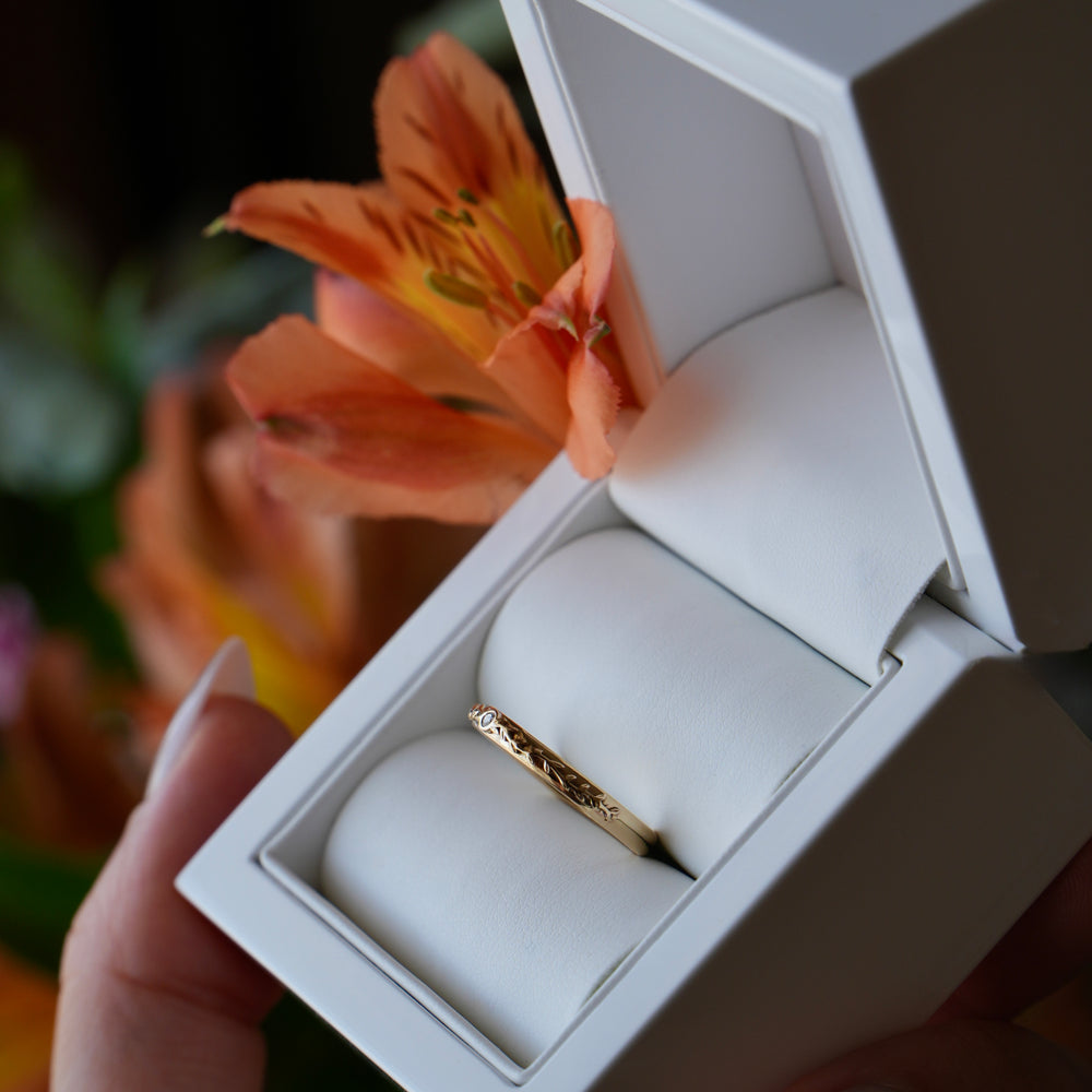 Gold ring in an open white jewelry box held by a hand with a blurred floral background