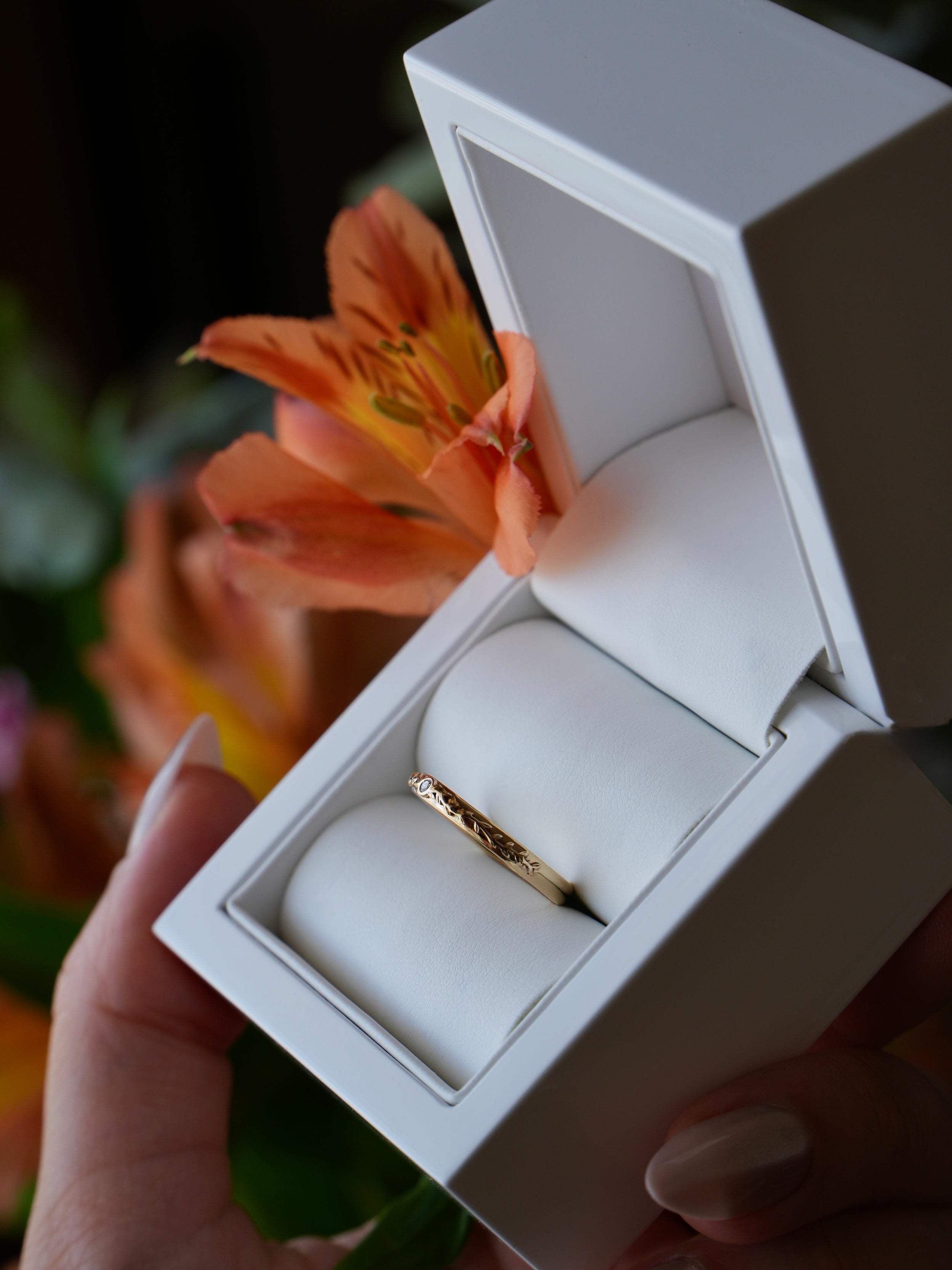 Gold ring in an open white jewelry box held by a hand with a blurred floral background