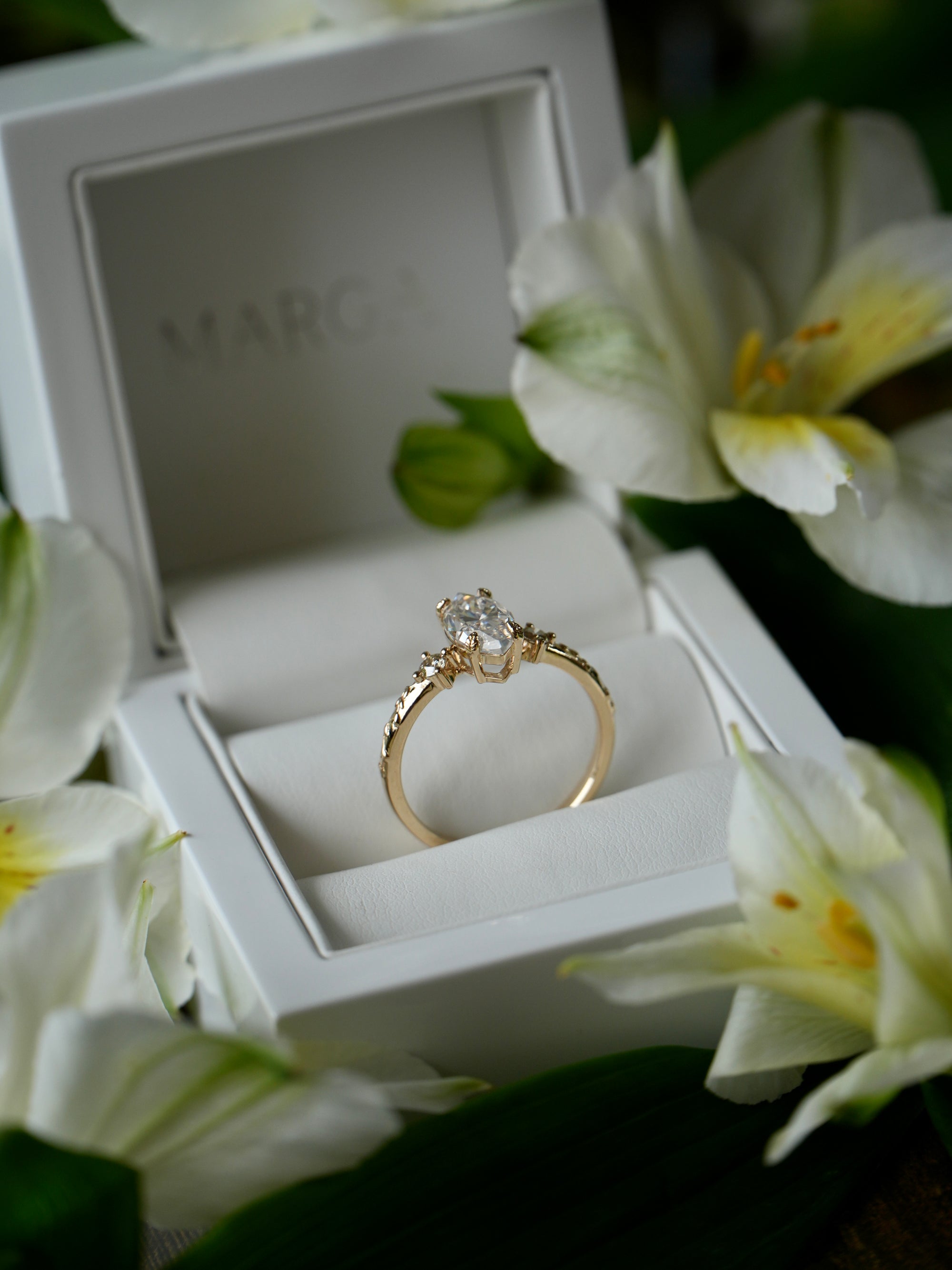 Gold ring with diamonds in a white jewelry box surrounded by white flowers