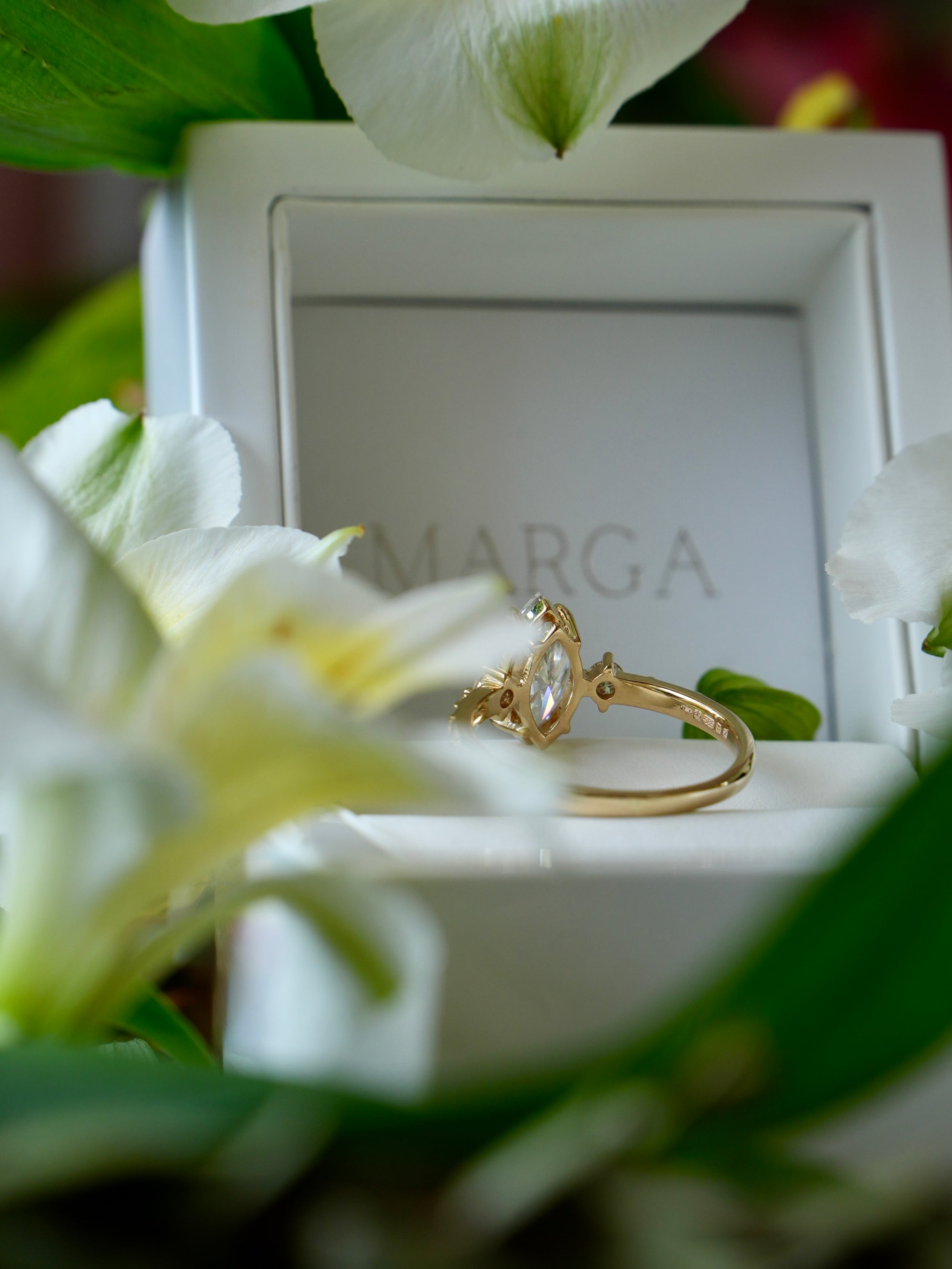 Gold ring in a white box with 'MARGA' branding, surrounded by white flowers.
