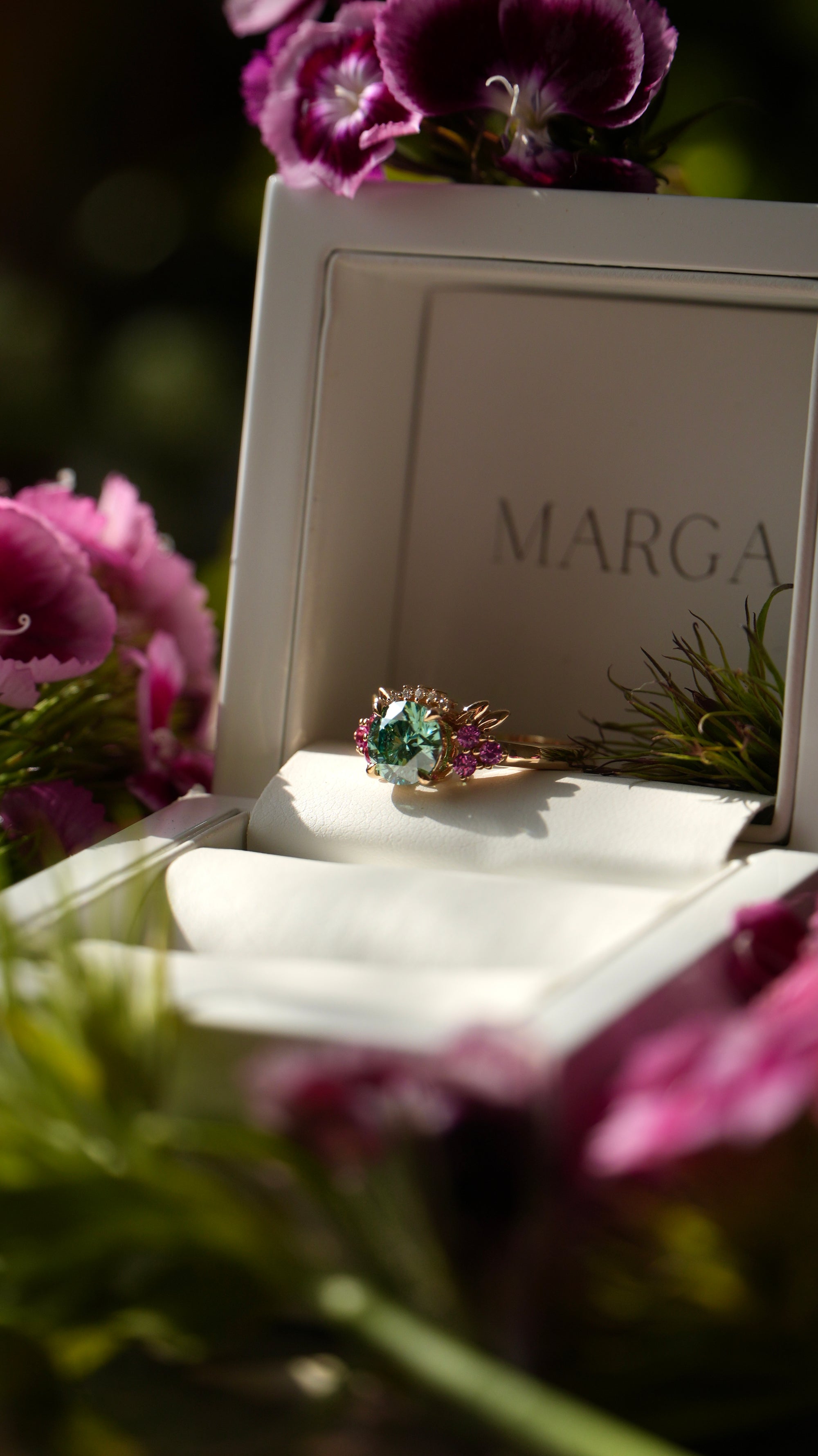 Ring in a jewelry box with 'MARGA' branding, surrounded by pink flowers.