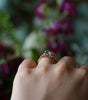 Hand wearing a ring with a green gemstone against a blurred floral background